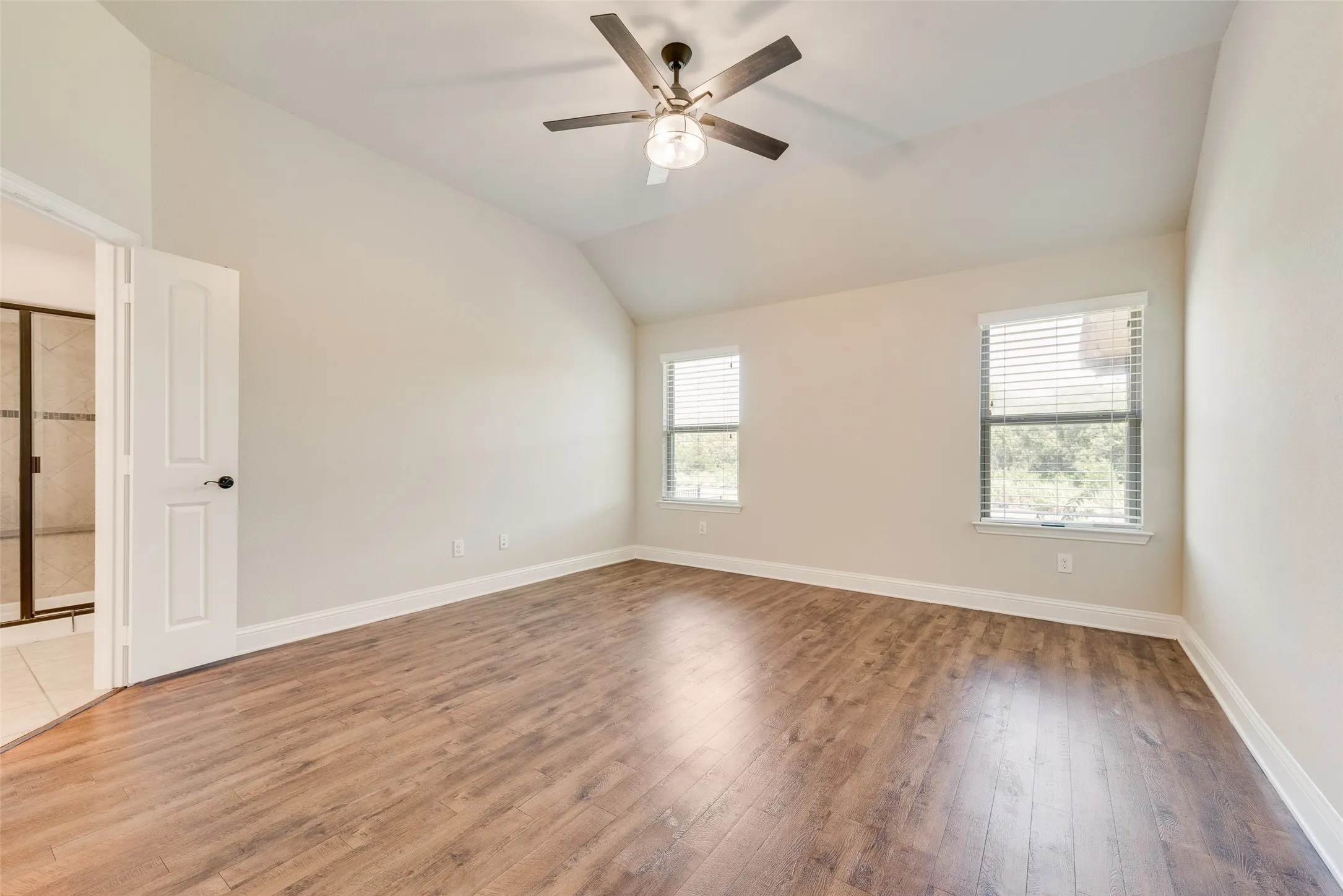 Vaulted ceiling with an attached ceiling fan, two windows with blinds, wood-finish flooring, and light-toned walls