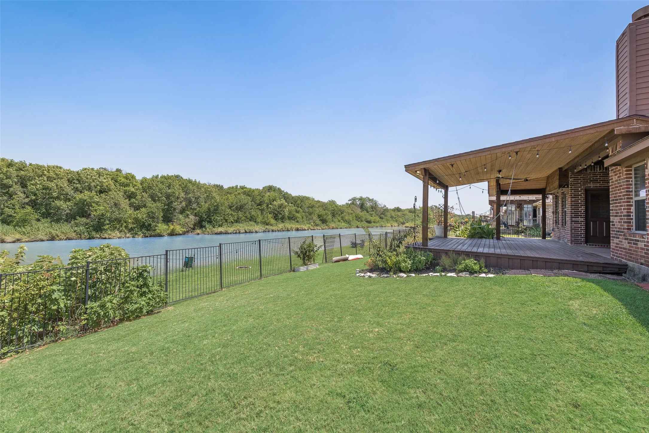 Expansive green lawn with a covered wood-finish deck, brick exterior, and a black metal fence bordering a natural waterway