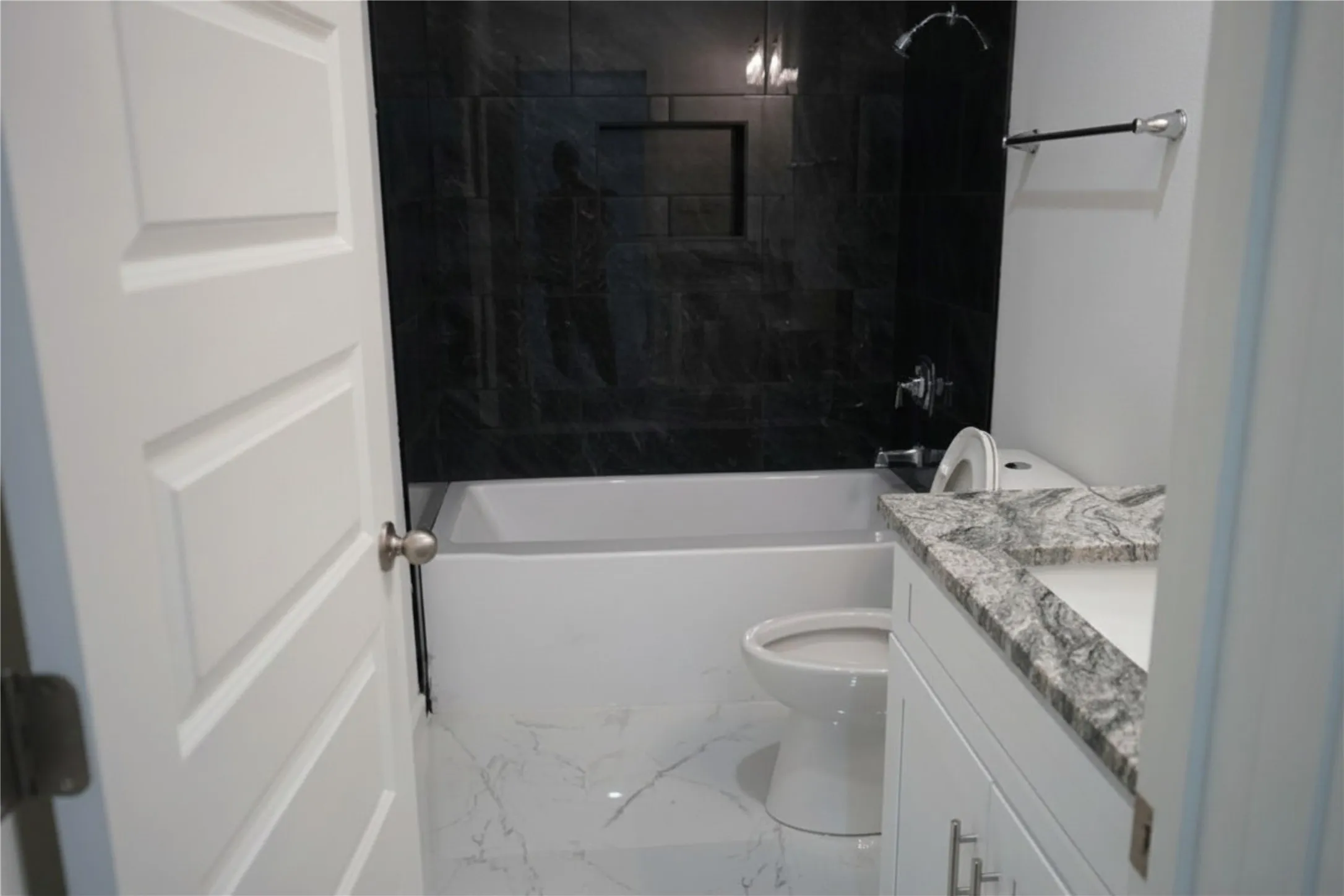 Bathroom featuring a white bathtub, dark tiled shower surround with a built-in niche, white vanity with a stone-look countertop, and a white toilet
