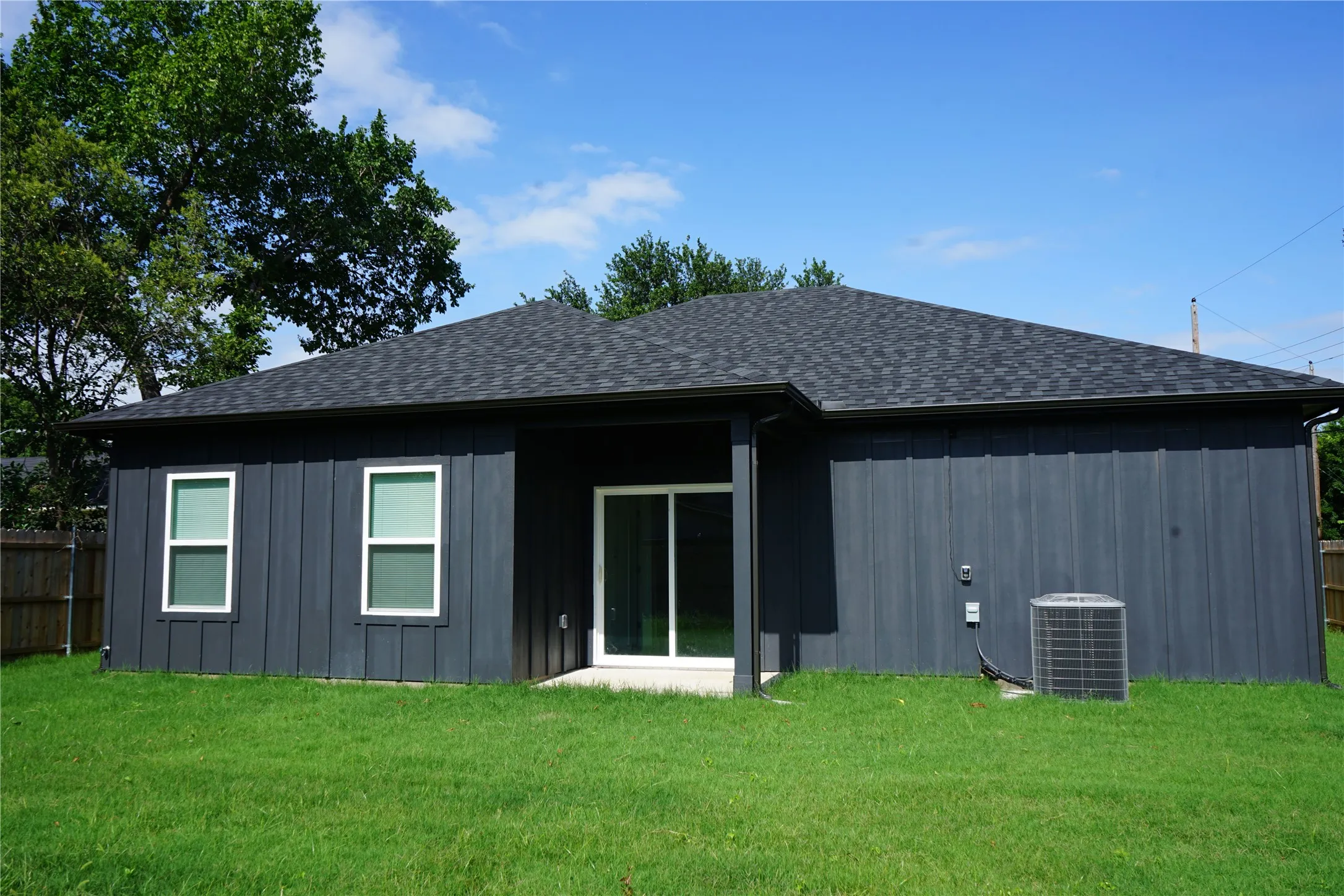 Dark gray vertical panel siding, dark asphalt shingle roof, white frame windows, sliding glass door, and a grass lawn