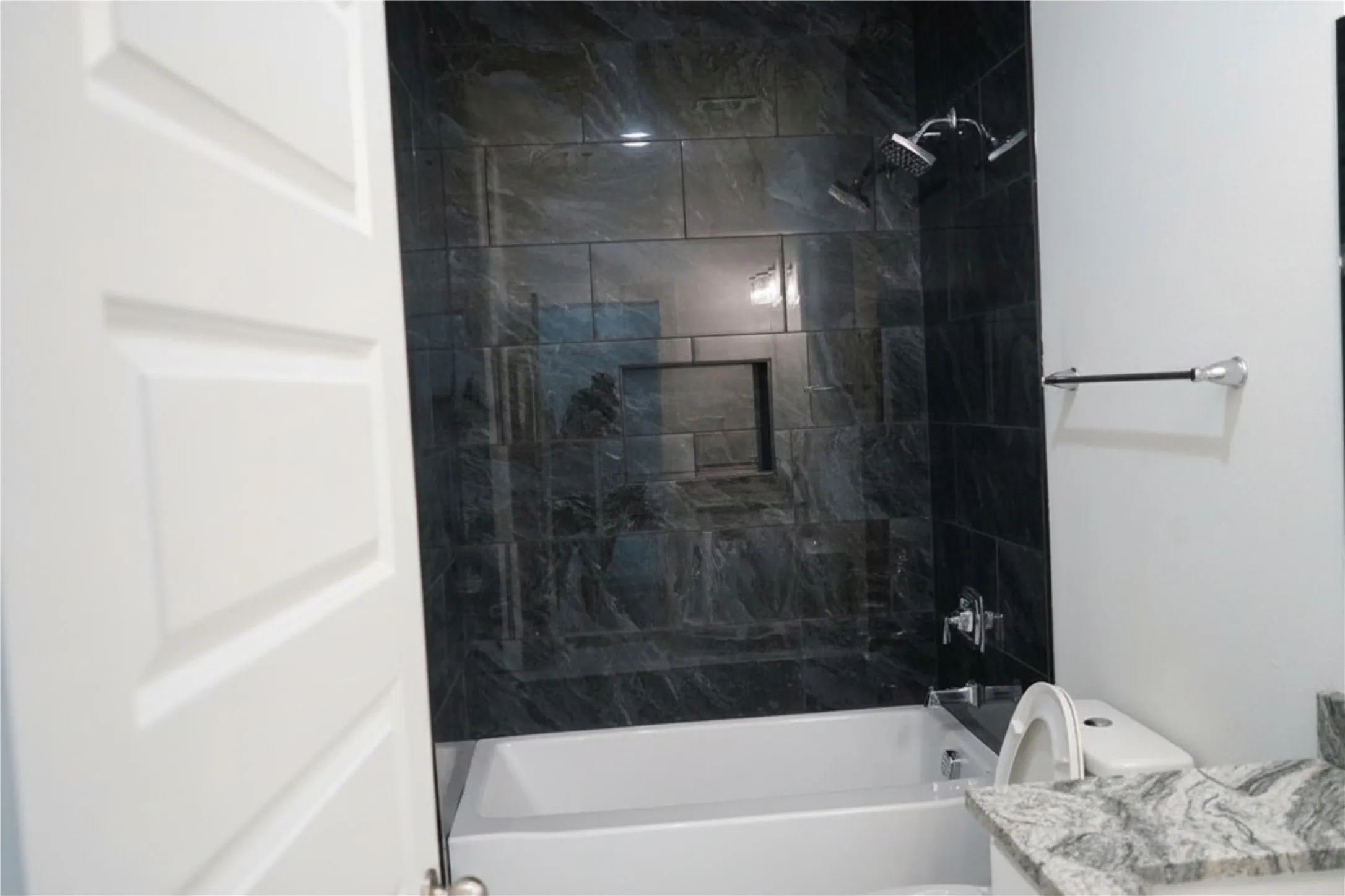 Bathtub with dark tile surround, built-in shower niche, dual shower heads, and an adjacent vanity with a stone countertop