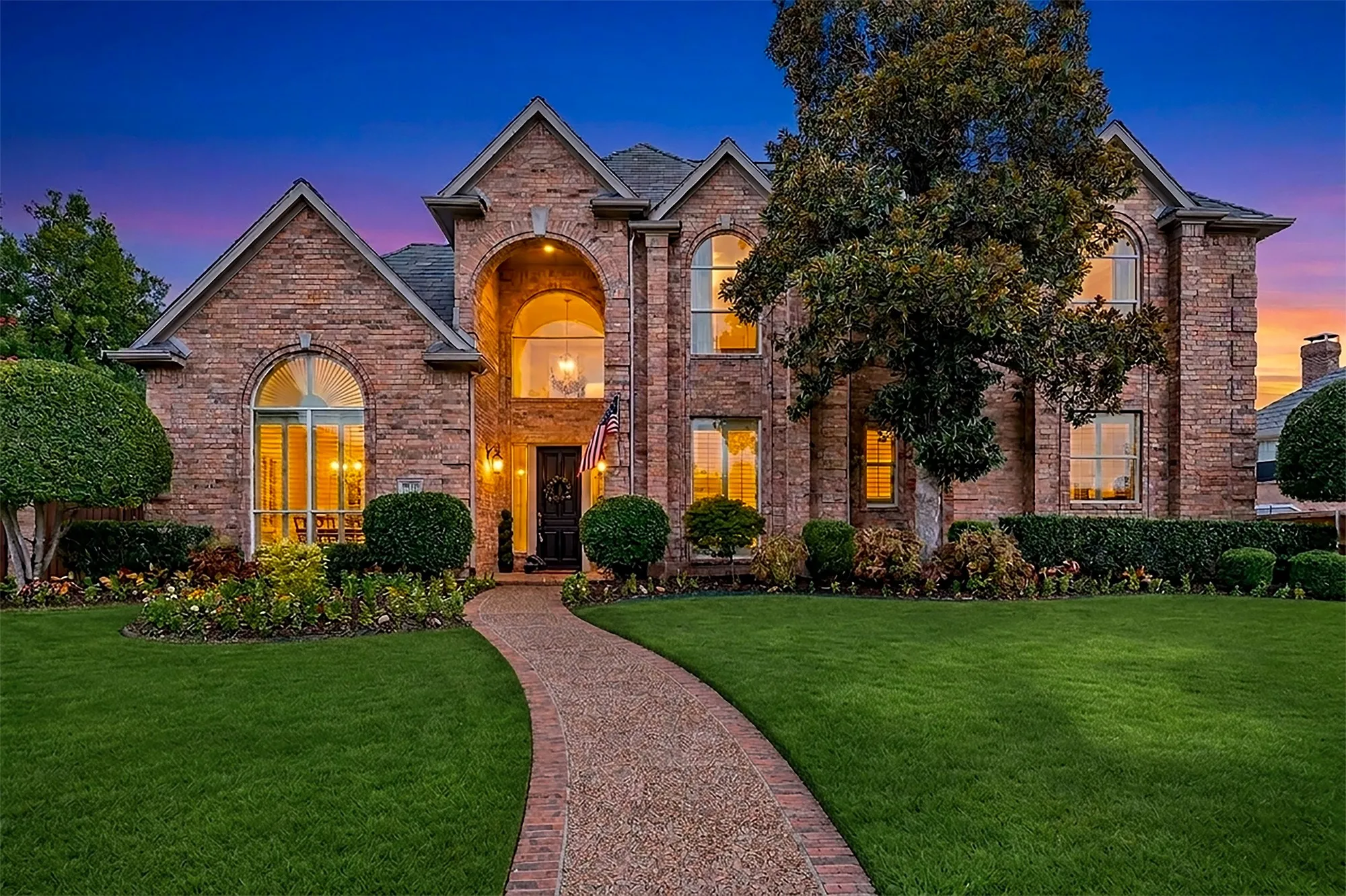 Welcome to 3616 Hearst Castle in Plano. A sweeping brick walkway guides toward a soaring double-height arched entryway lined with manicured lawn landscaping. Step home each evening greeted by warm, golden window light glowing throughout every room