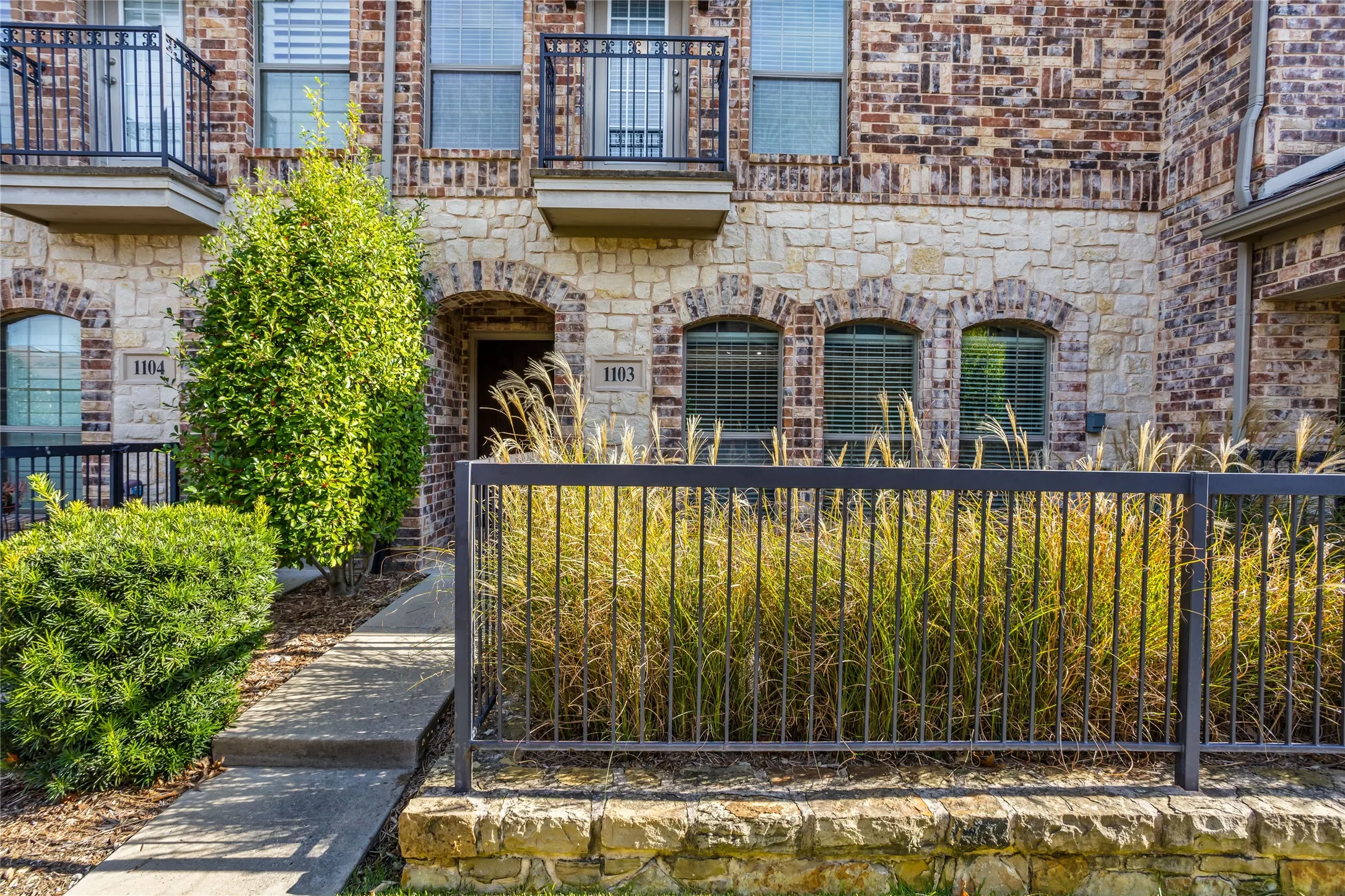 Brick and stone facade featuring arched windows, Juliet balconies with wrought iron railings, and a concrete pathway with mature landscaping