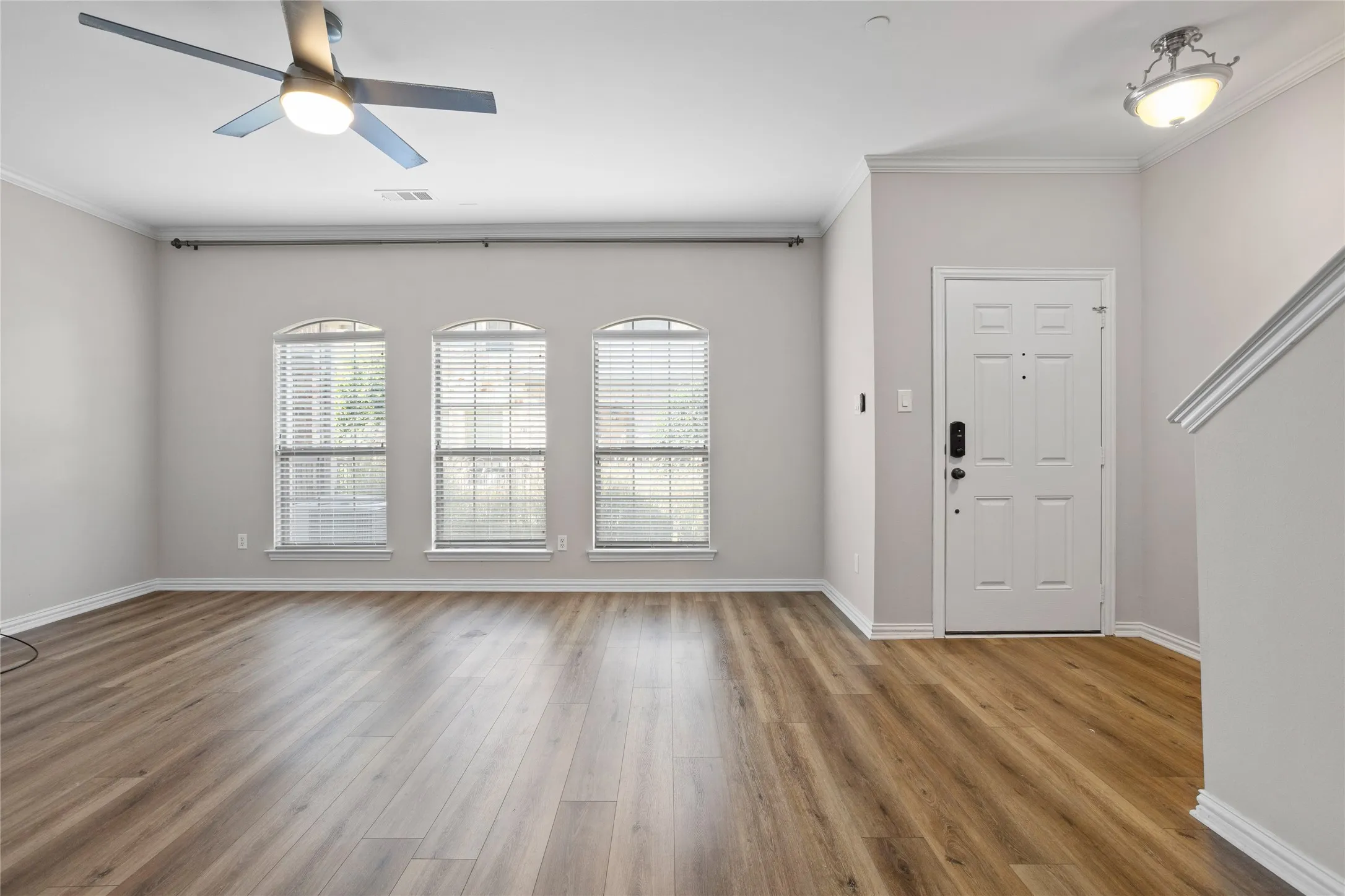 Wood-finish flooring, three arched windows with blinds, ceiling fan, white paneled door, and built-in ceiling light fixture