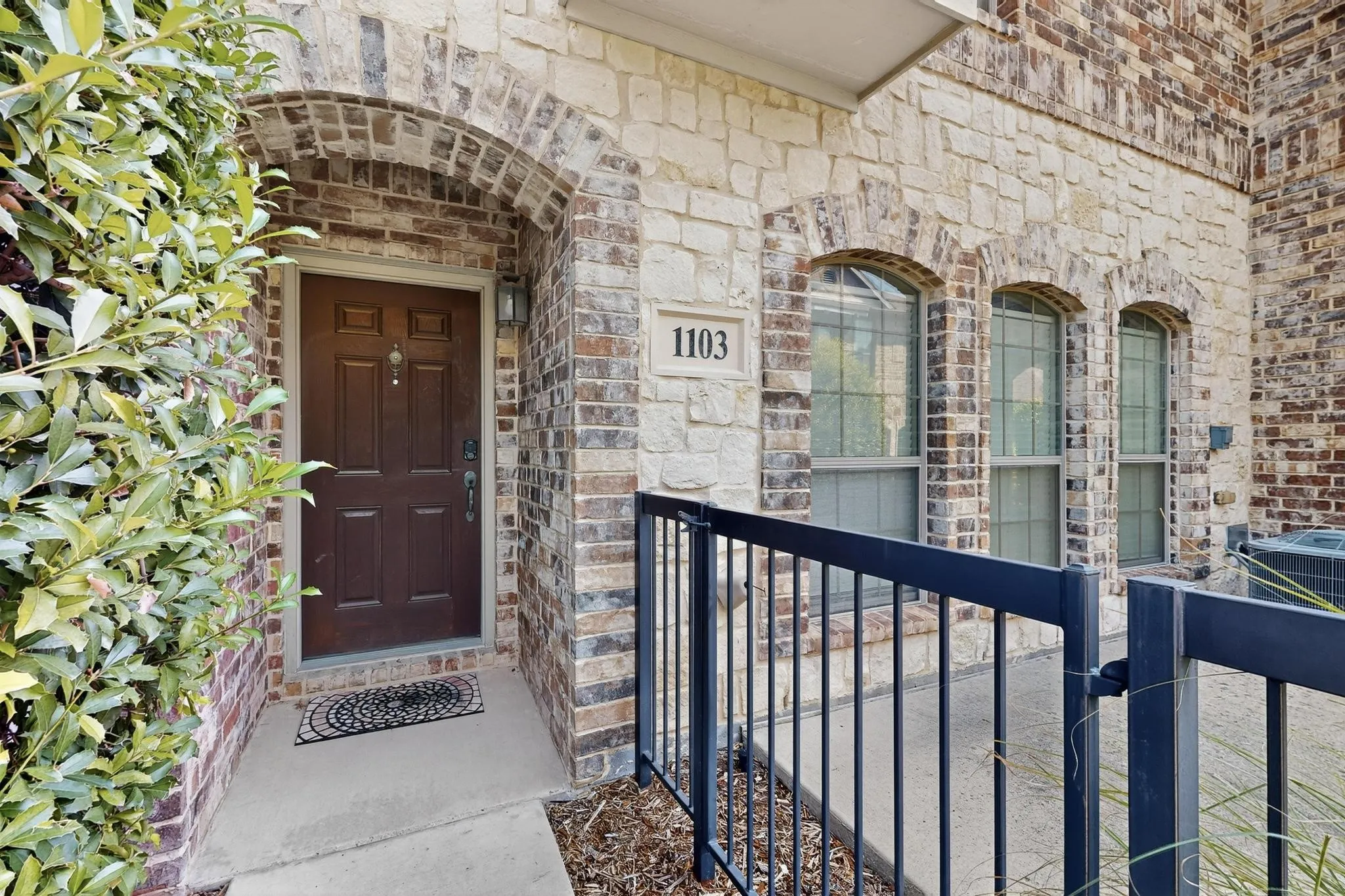 Brick and stone facade featuring arched window details, a dark wood-finish entry door, and a black metal railing