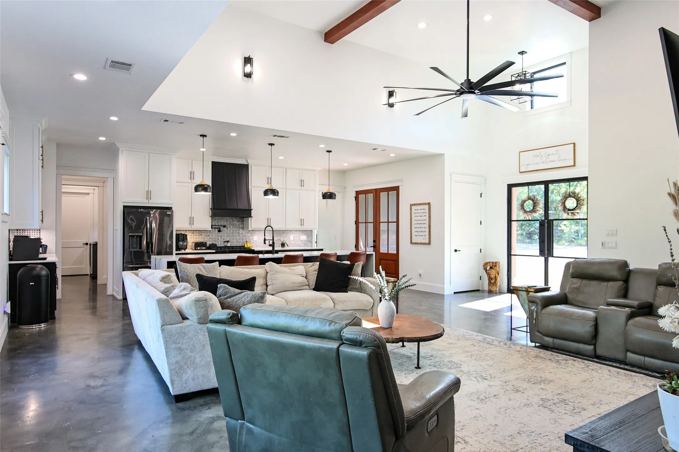 Vaulted ceiling with exposed wood beams, polished concrete flooring, white shaker cabinetry, large kitchen island with pendant lighting, and black framed glass doors
