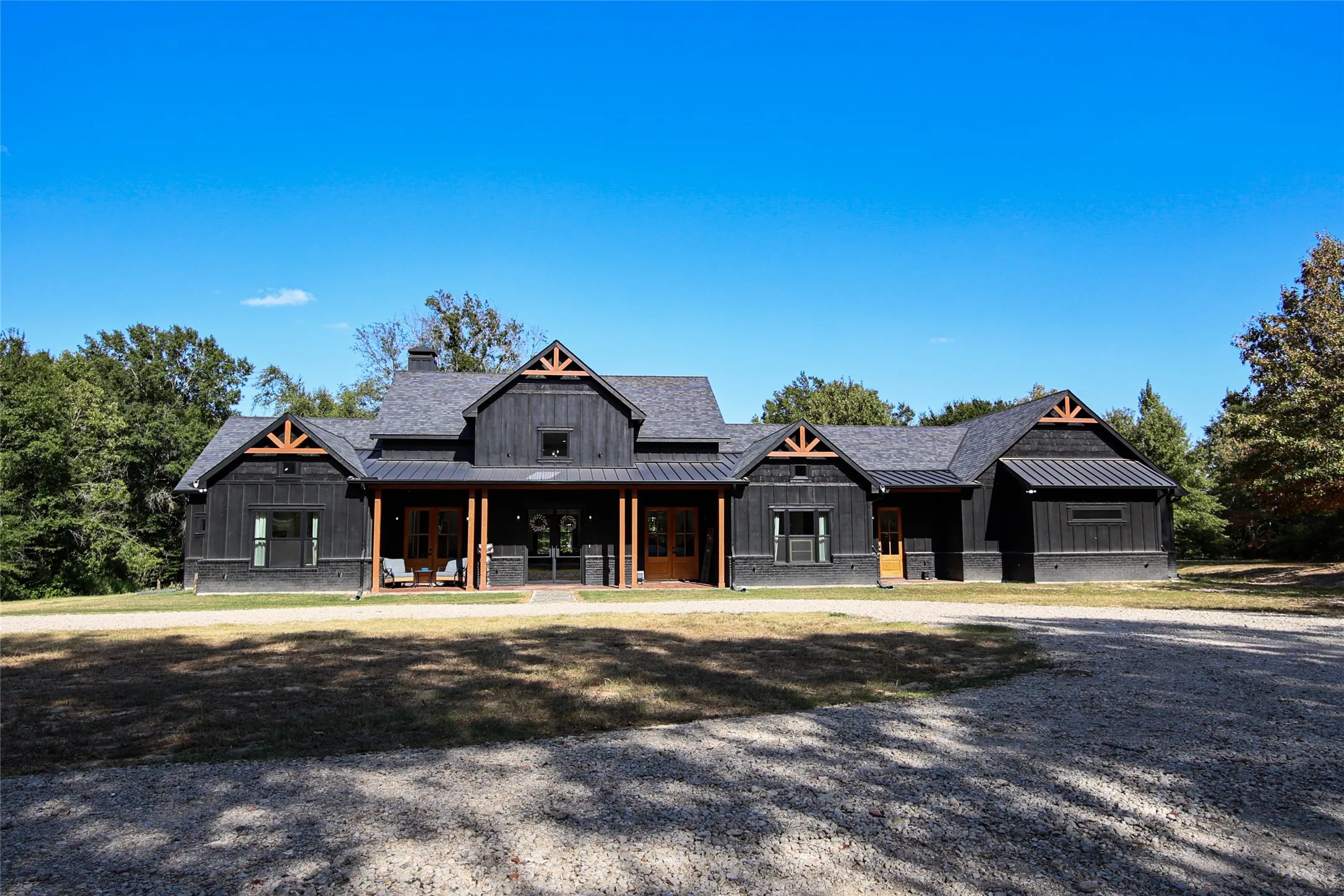 Modern farmhouse exterior featuring black board and batten siding, a covered porch with timber columns, exposed timber gable trusses, a shingle roof, and a gravel driveway