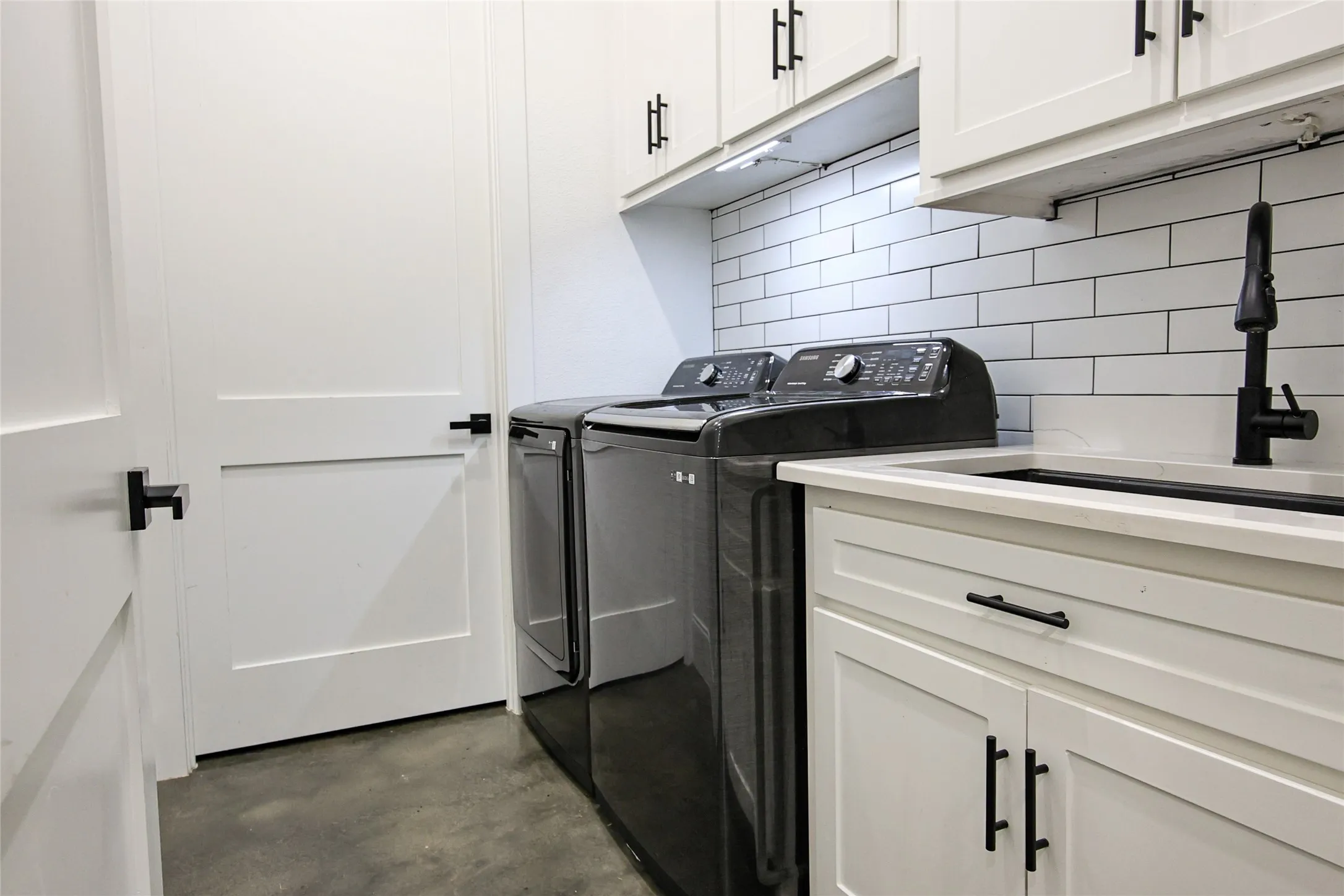 White shaker-style cabinetry with matte black hardware, integrated utility sink, subway tile backsplash, and concrete-finish flooring