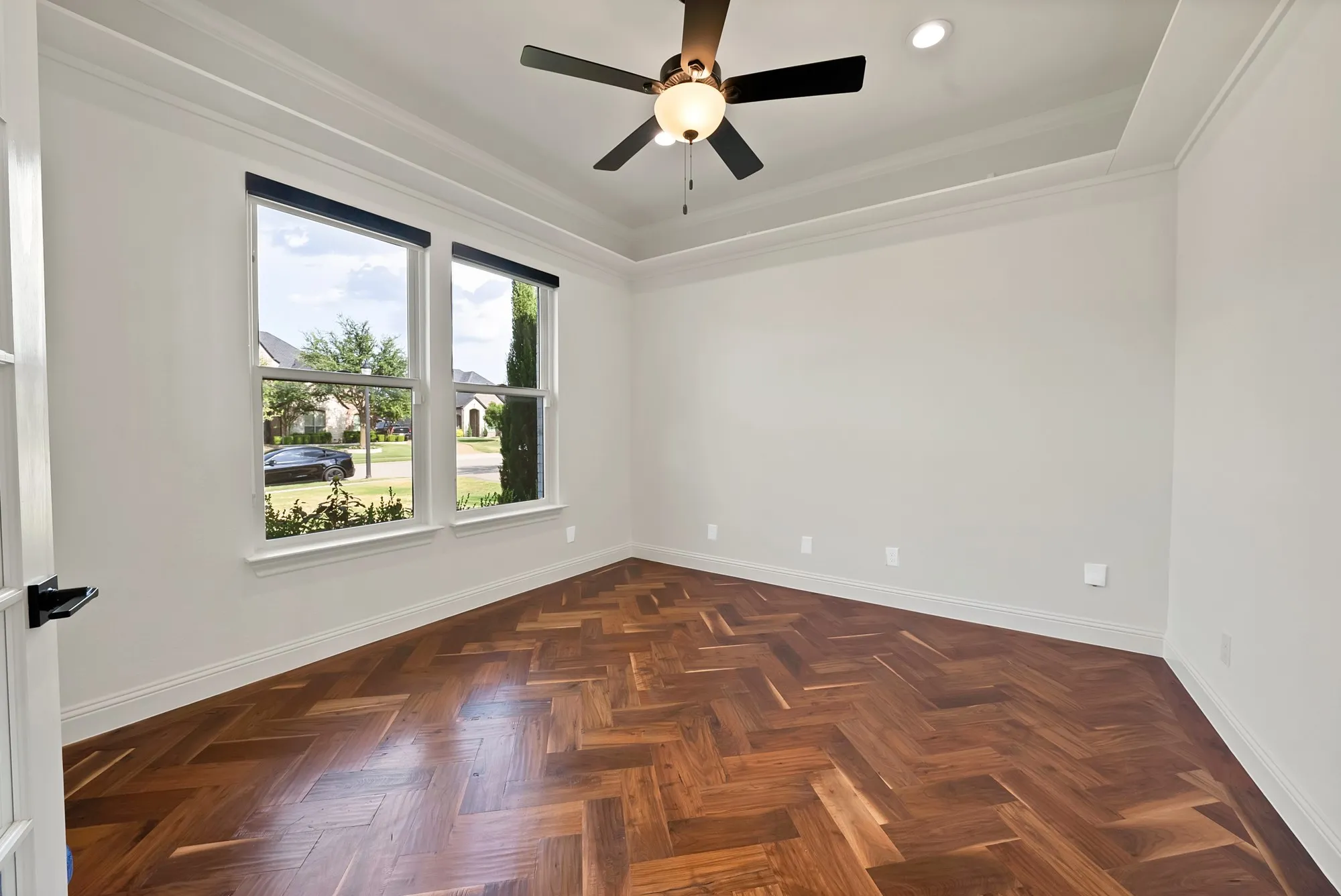 Bright corner bedroom showcasing herringbone wood-finish flooring, ceiling fan with integrated lighting, and large double-hung windows framing natural light and outdoor scenery