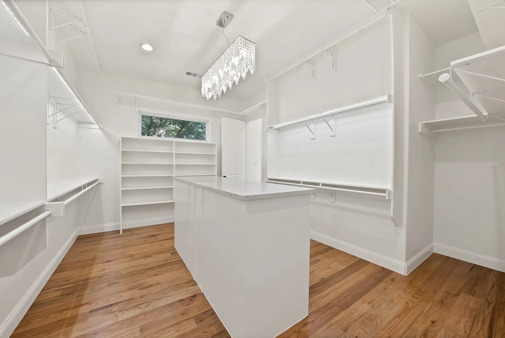 Wood-finish flooring, central island with solid surface countertop, built-in shelving, white walls, and a contemporary chandelier