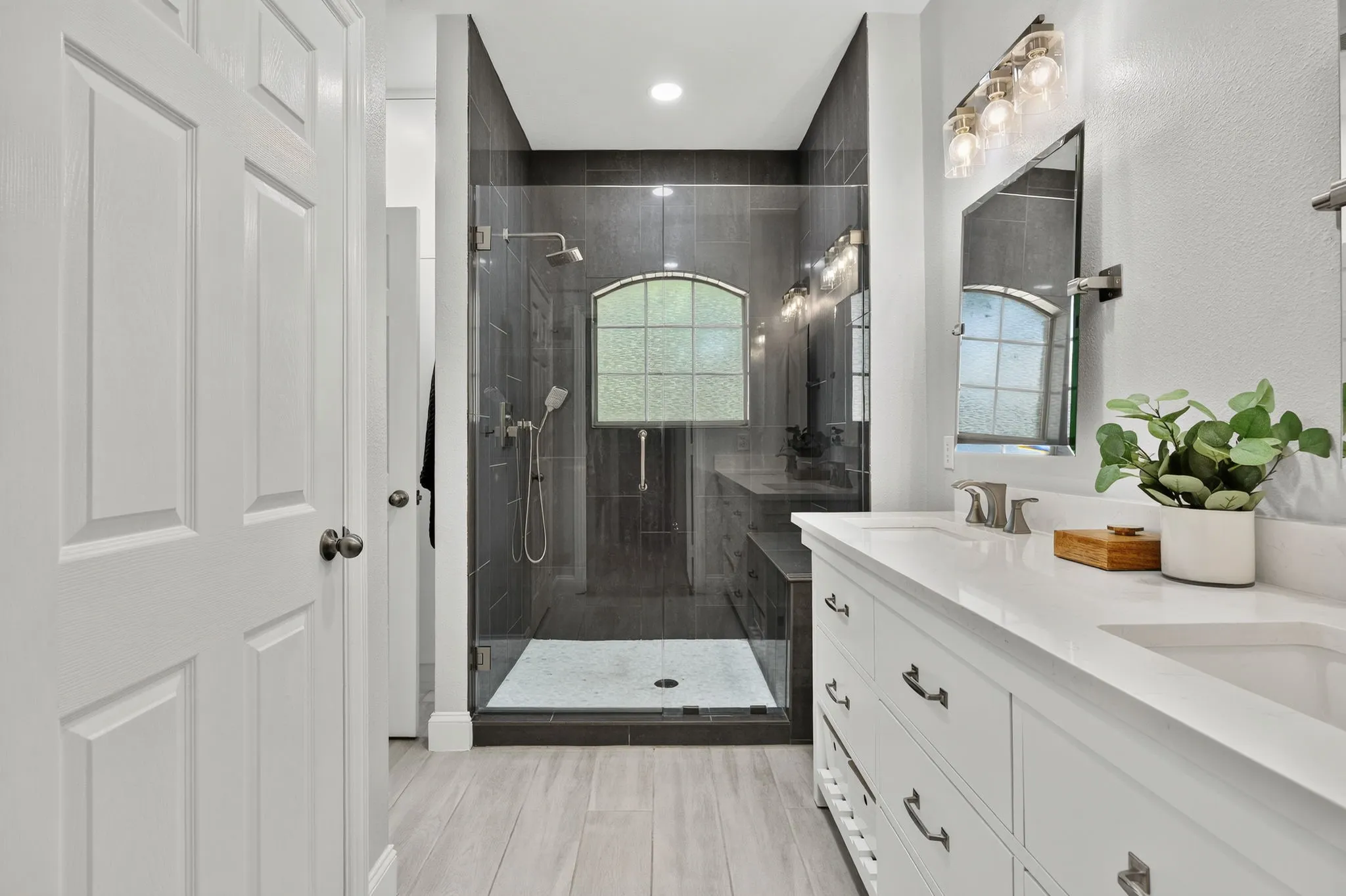 Dual vanity with quartz countertop, walk-in shower with tiled walls, glass shower enclosure, and wood-finish flooring