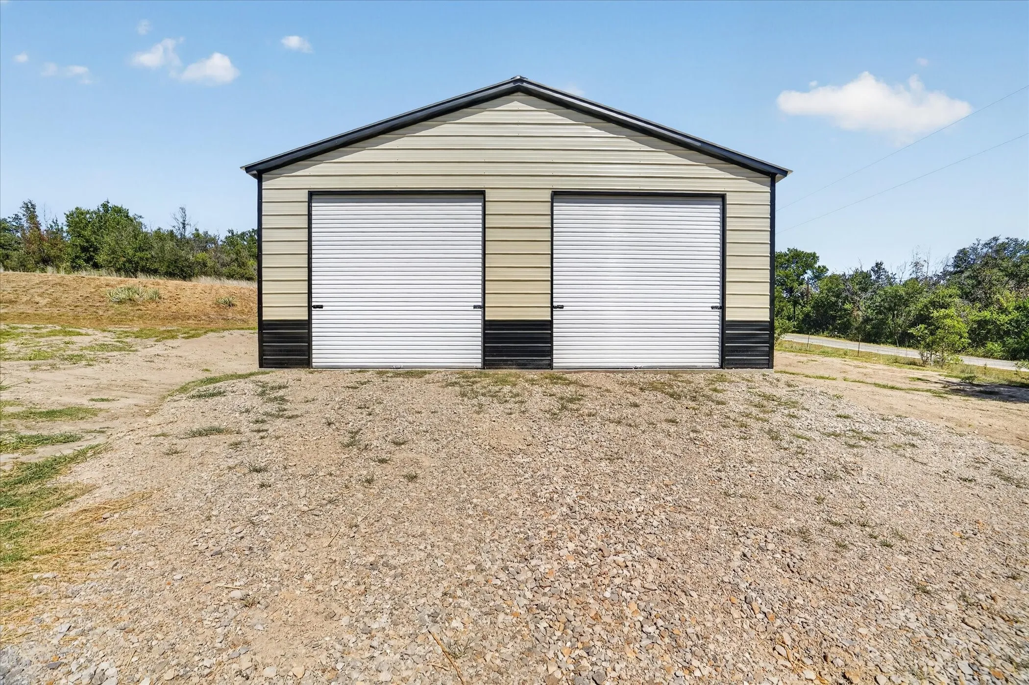 Two-car metal garage with a gabled roof and roll-up doors, set on a gravel surface