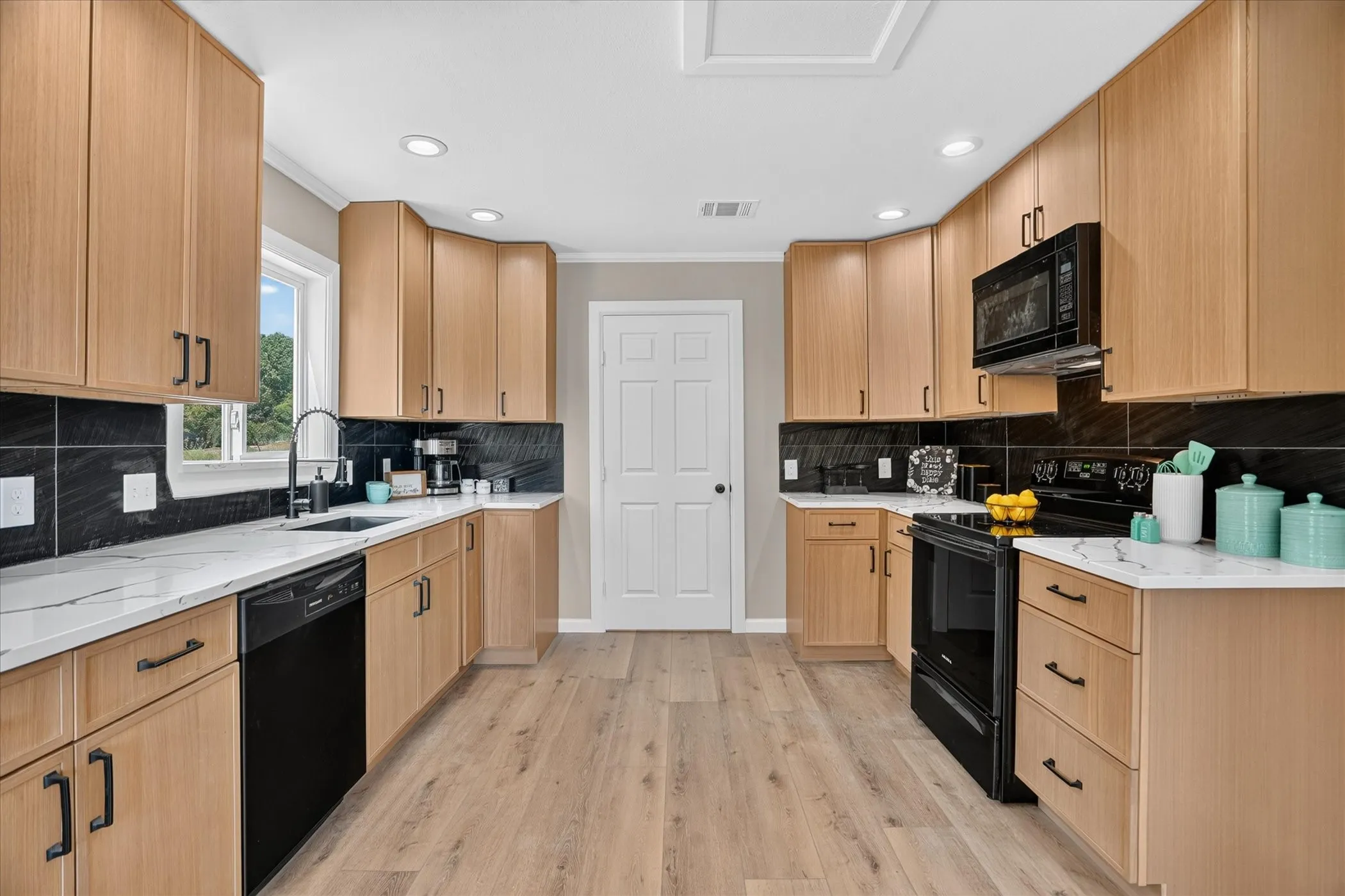 Kitchen featuring light wood-finish cabinetry, black tile backsplash, white countertops, stainless steel sink with a matte black faucet, and wood-finish flooring