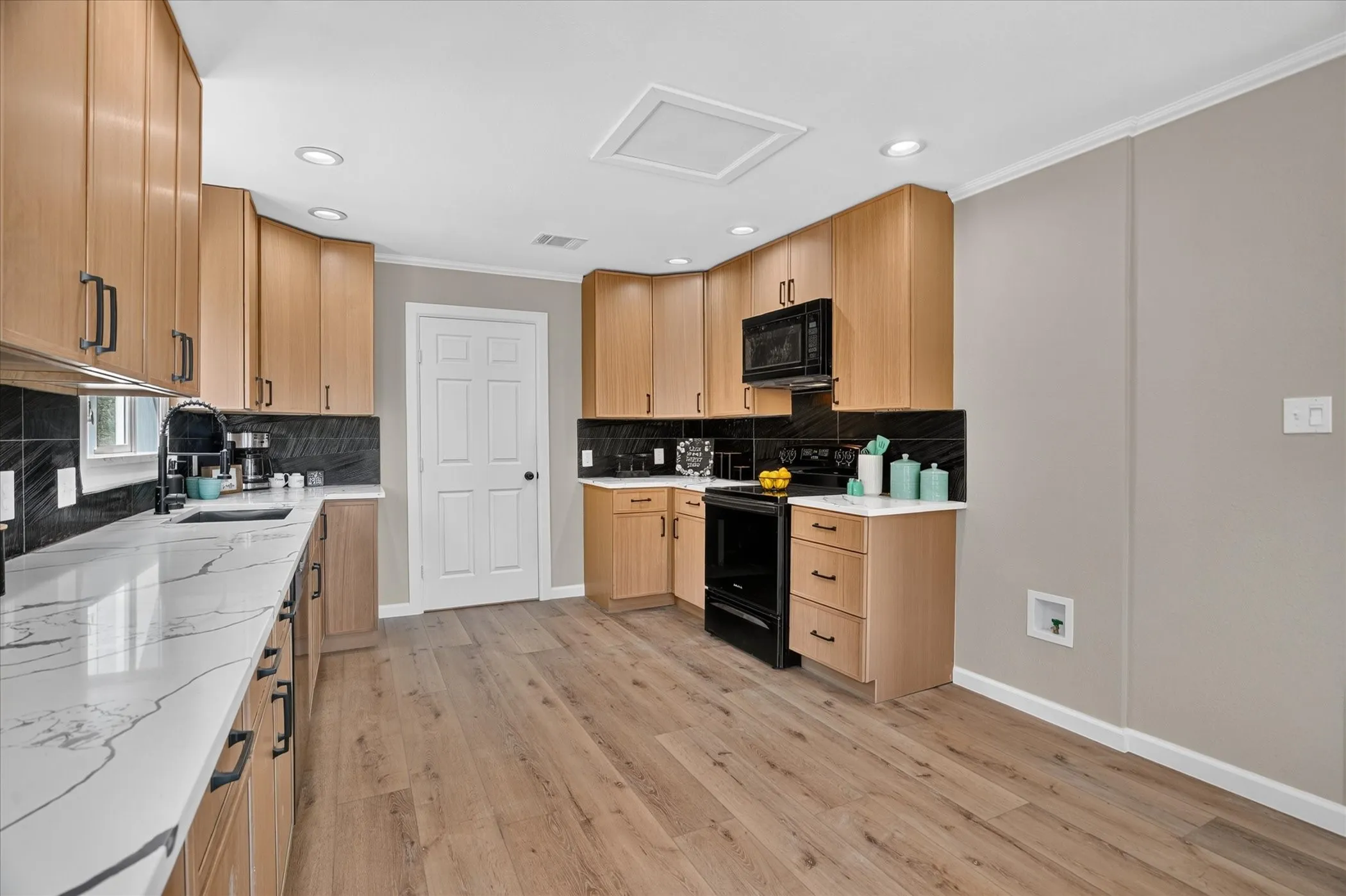 Kitchen featuring wood-finish flooring, light wood cabinetry, black hardware, black tile backsplash, and white countertops