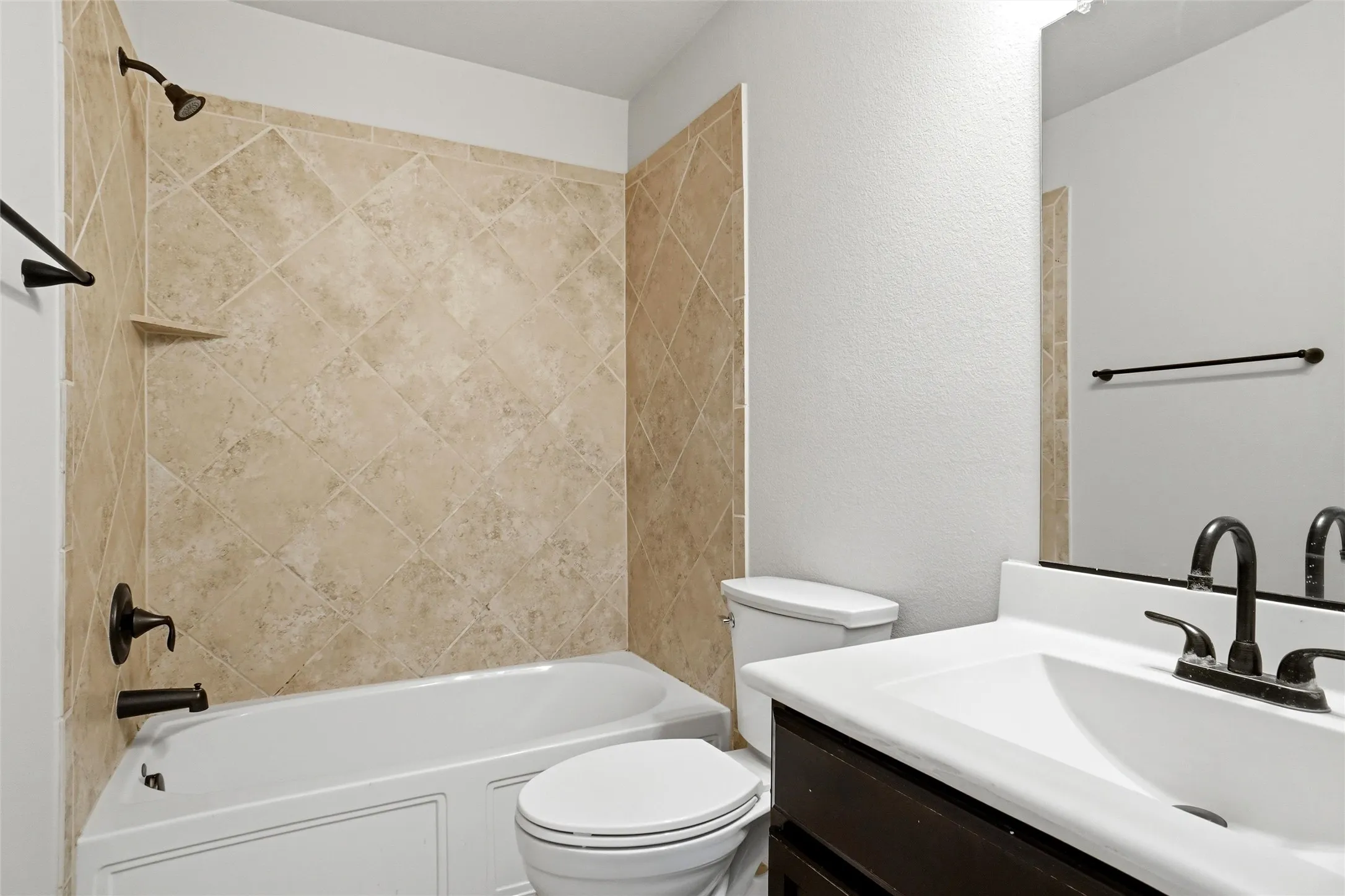 Bathtub with tiled surround, dark bronze fixtures, white vanity with integrated sink, and dark wood-finish cabinetry