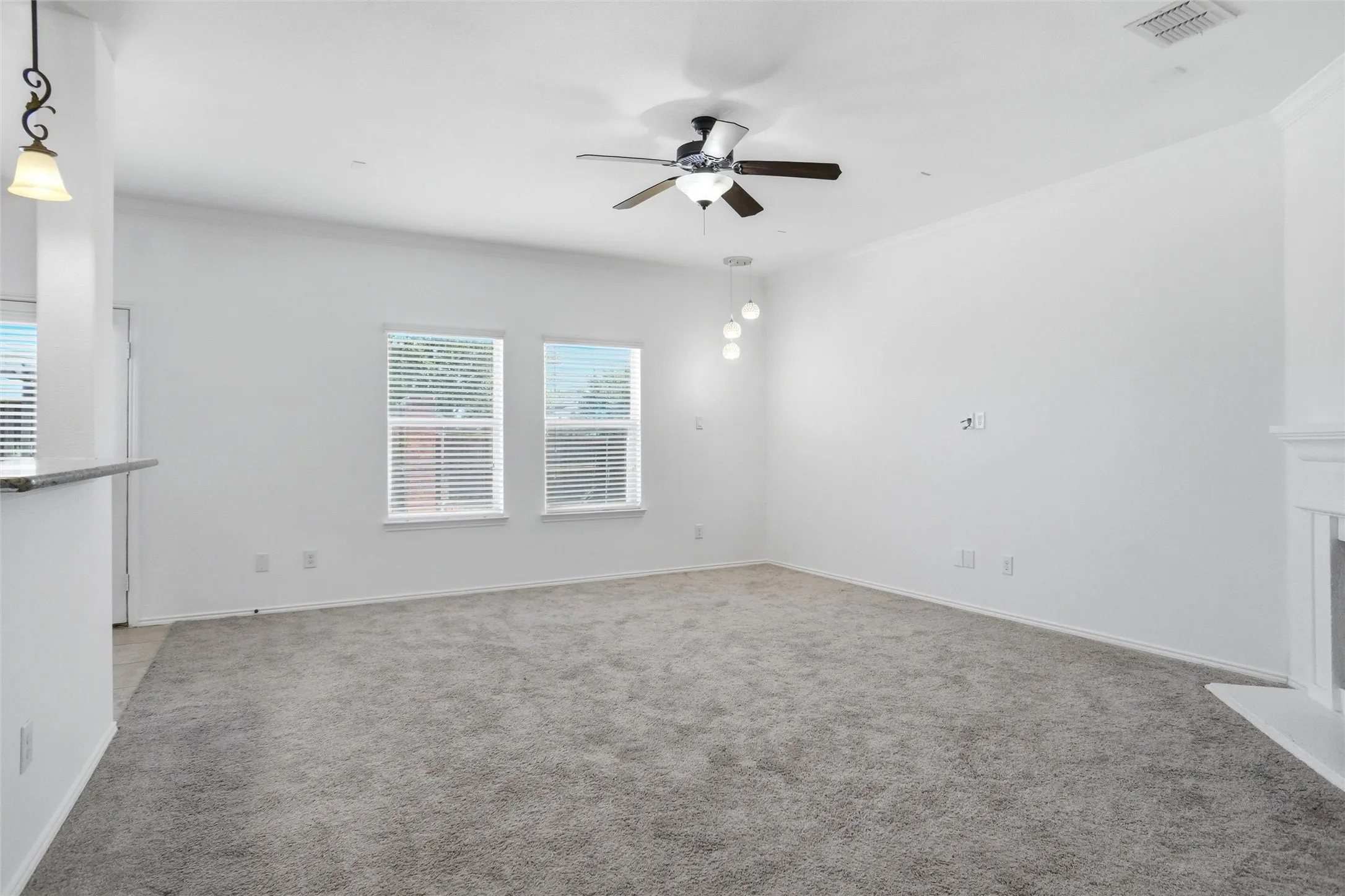 Spacious room featuring carpeting, two windows with blinds, a ceiling fan, a pendant light fixture, and a fireplace surround