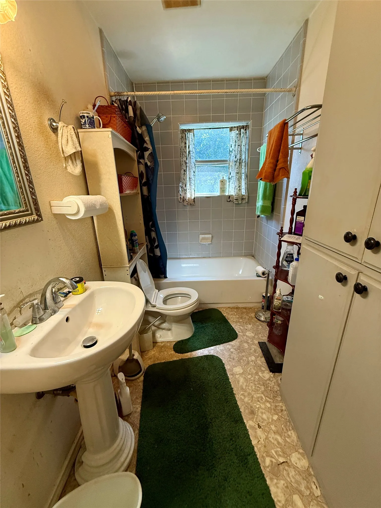 Pedestal sink, built-in bathtub, light-toned wall tiling, window, and cabinet storage