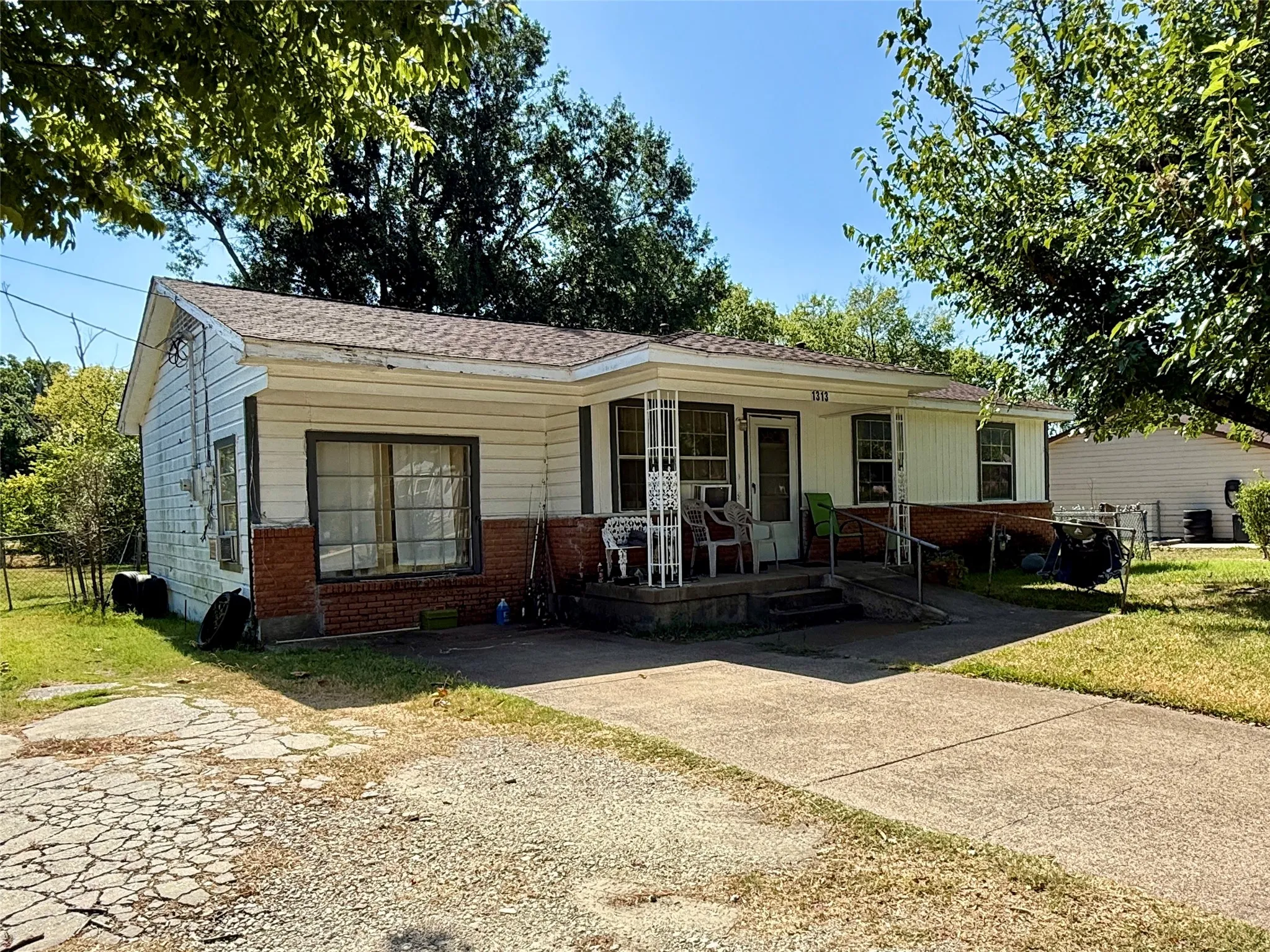 White siding exterior with a brick foundation, a covered front porch, a concrete driveway, and mature trees