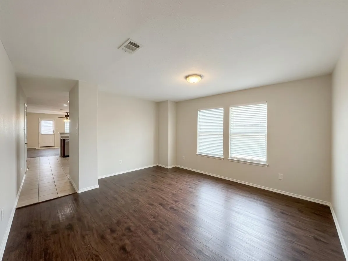 Spacious room featuring wood-finish flooring, light-colored walls, dual windows with blinds, and a ceiling-mounted light fixture