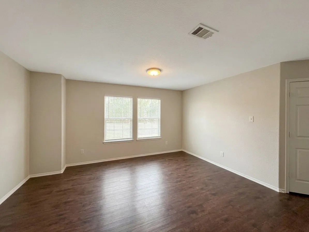 Spacious room featuring wood-finish flooring, neutral-toned walls, multiple windows with blinds, and a flush-mount ceiling light