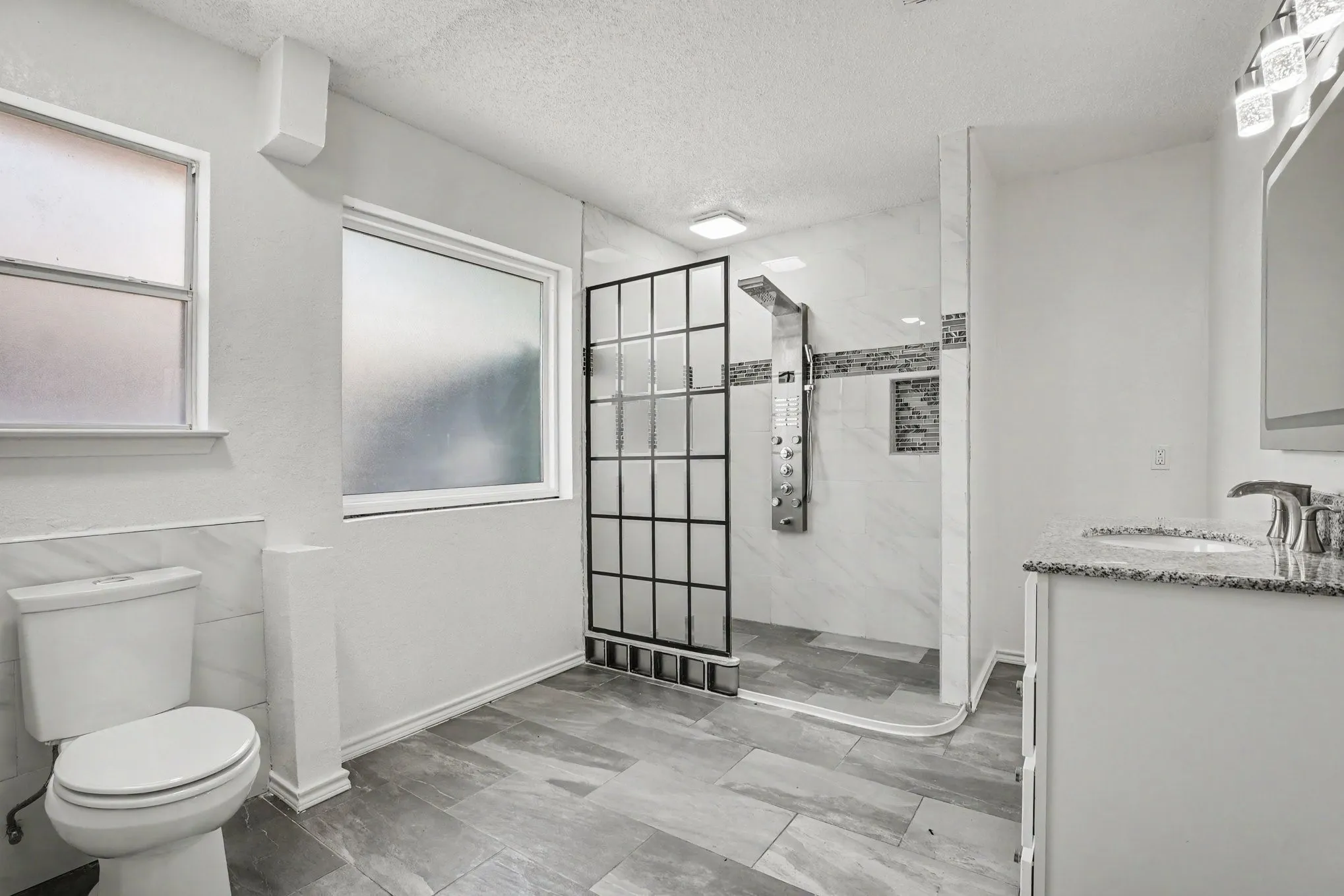 Walk-in shower with grid-patterned glass enclosure, vanity with granite-style countertop, modern light fixture, and tile flooring
