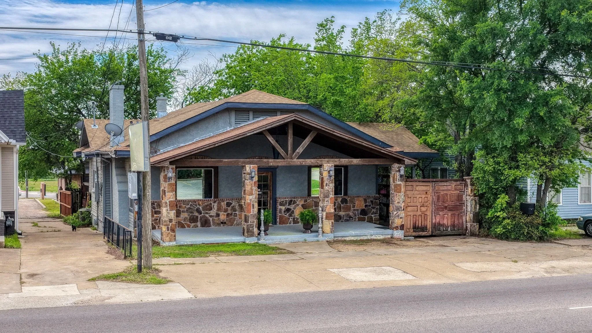 Stone veneer columns supporting a pitched porch roof with exposed wooden beams, a gray facade, and a gated side entry