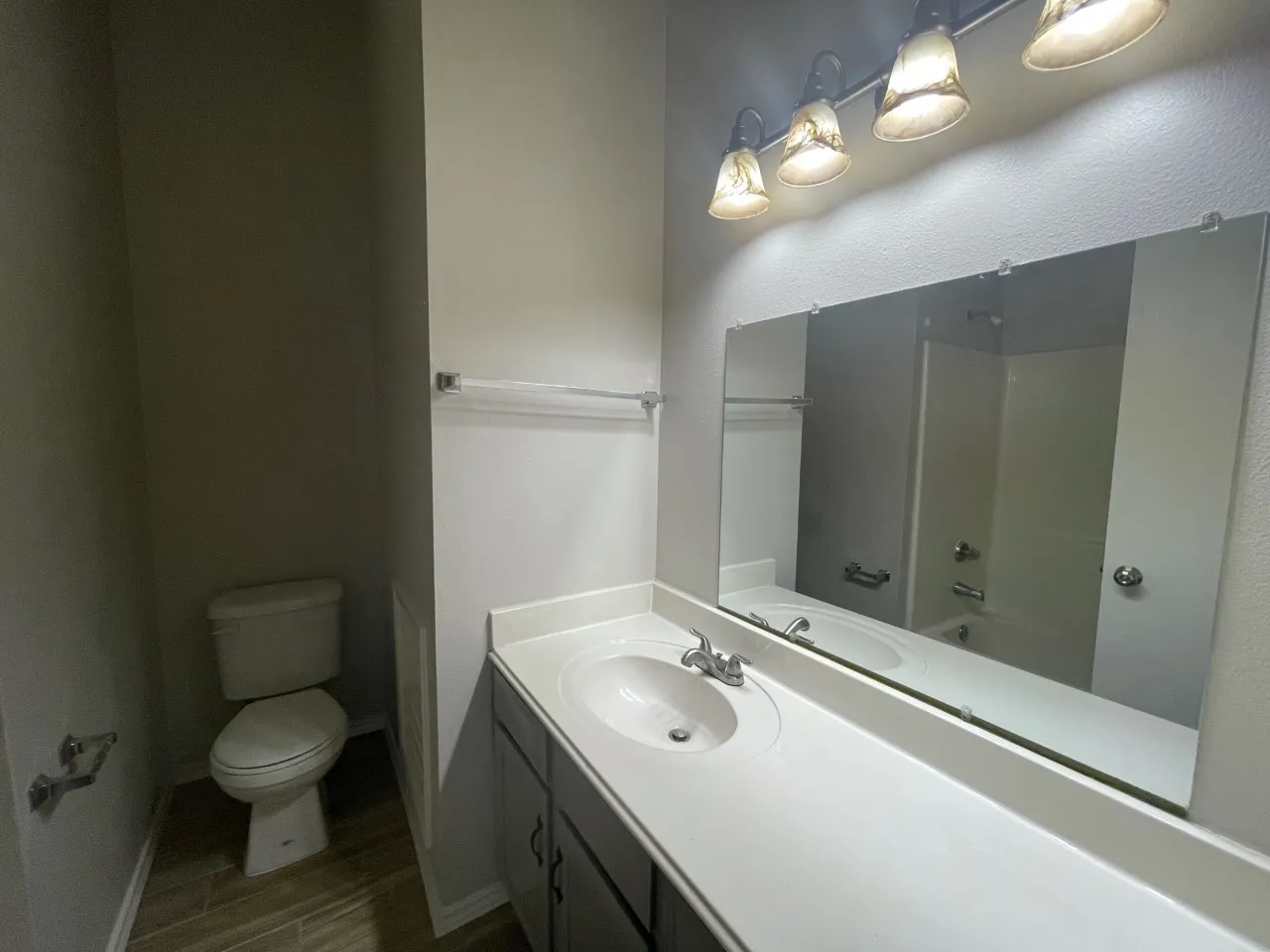 Vanity with integrated sink and chrome faucet, large wall-mounted mirror, four-light vanity fixture, toilet, and wood-finish tile flooring