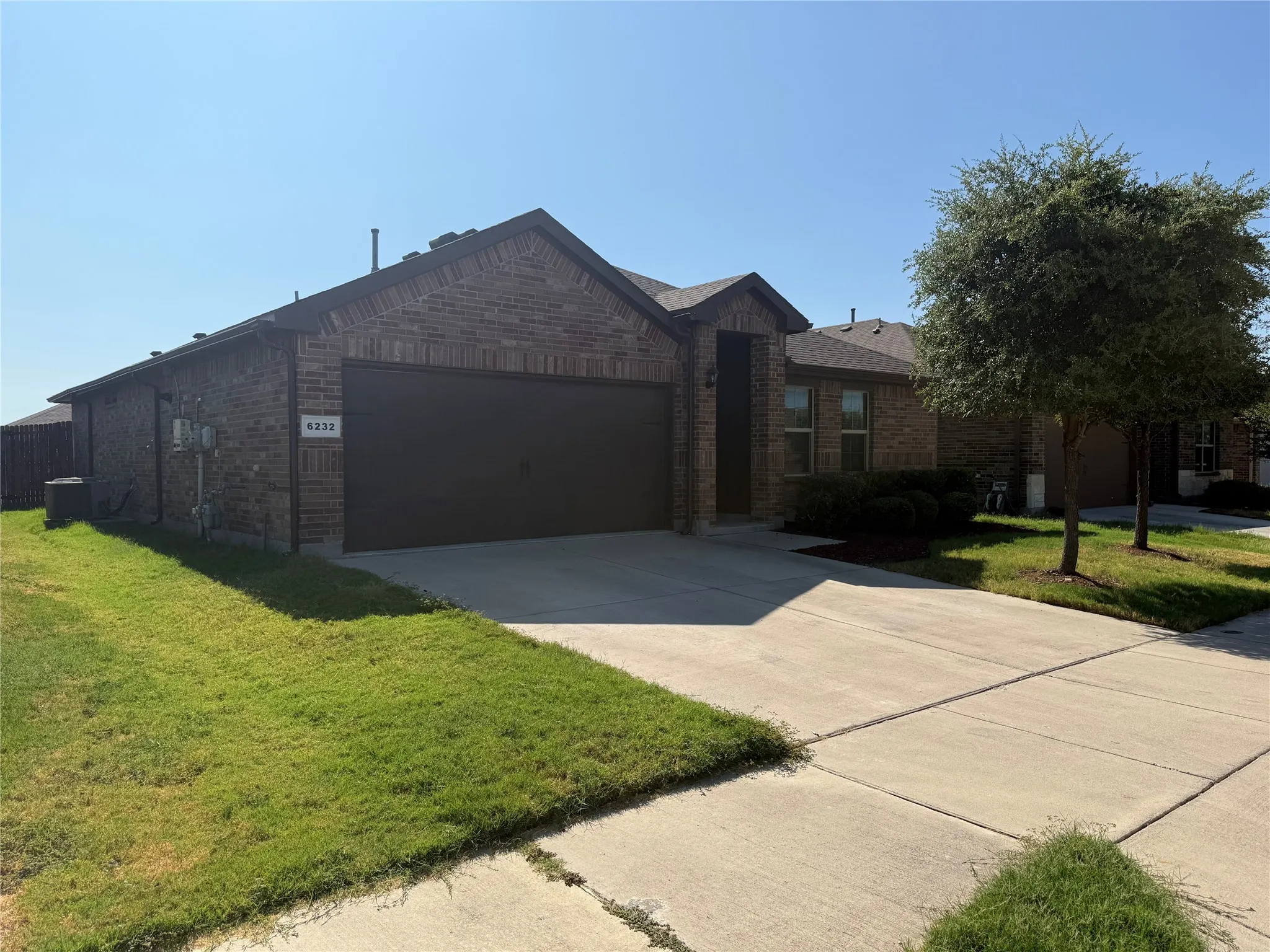 Brick facade with dark trim, attached garage, concrete driveway, and established landscape trees