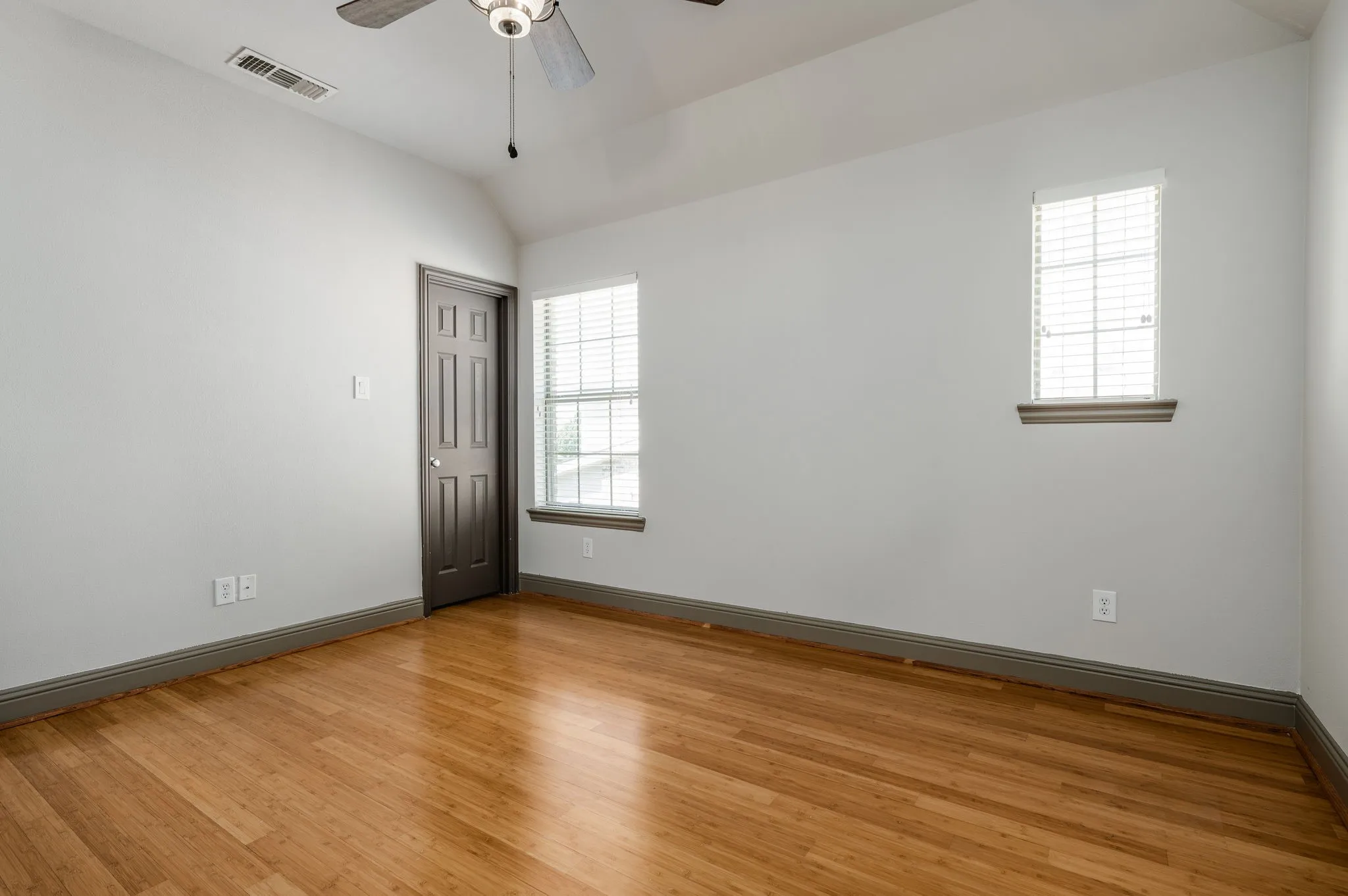Hardwood-style flooring, vaulted ceiling, ceiling fan, two windows with blinds, and a paneled door