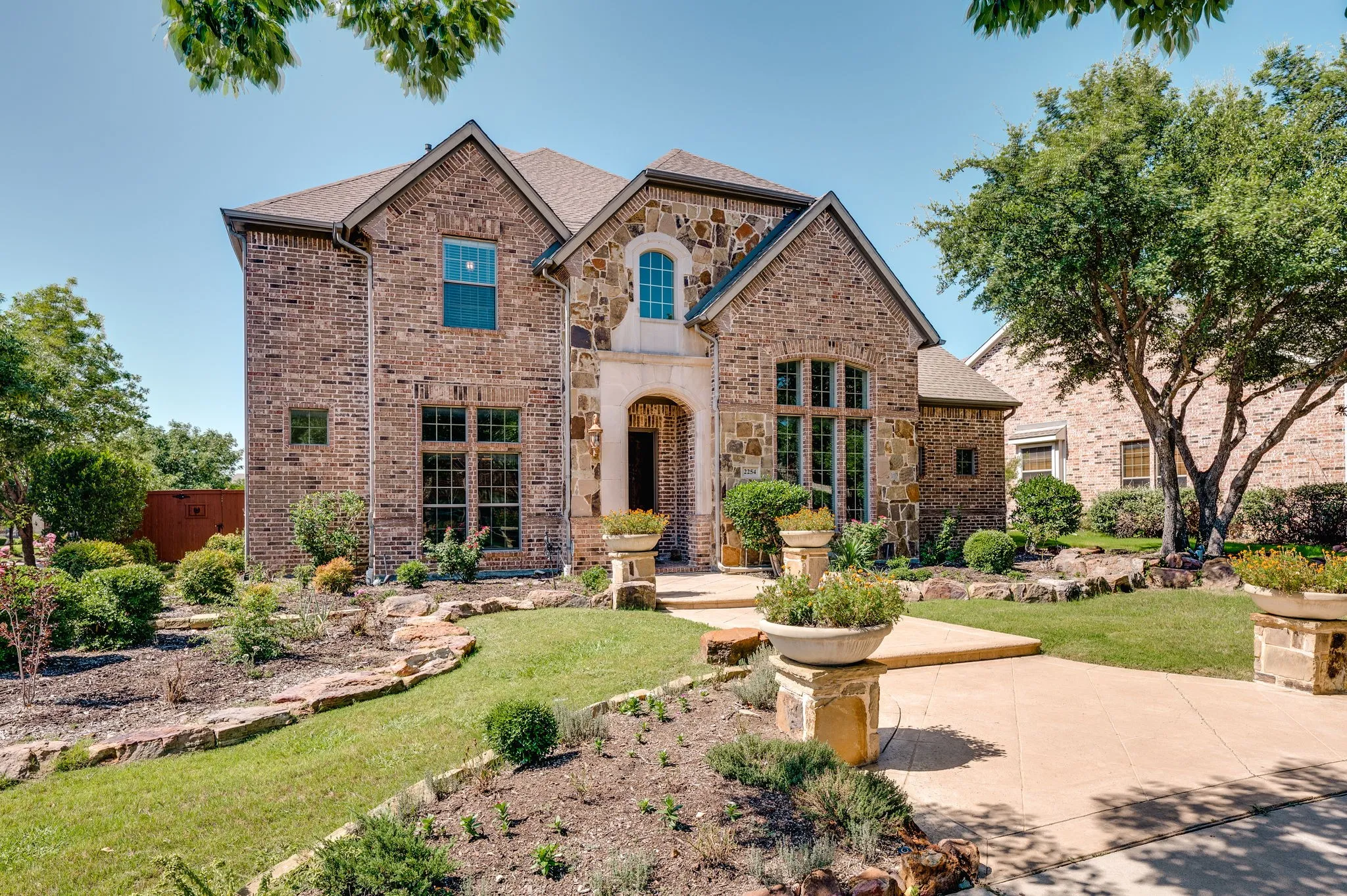 Brick and stone facade, arched entryway, gabled roofline, mature trees, and tiered landscaping