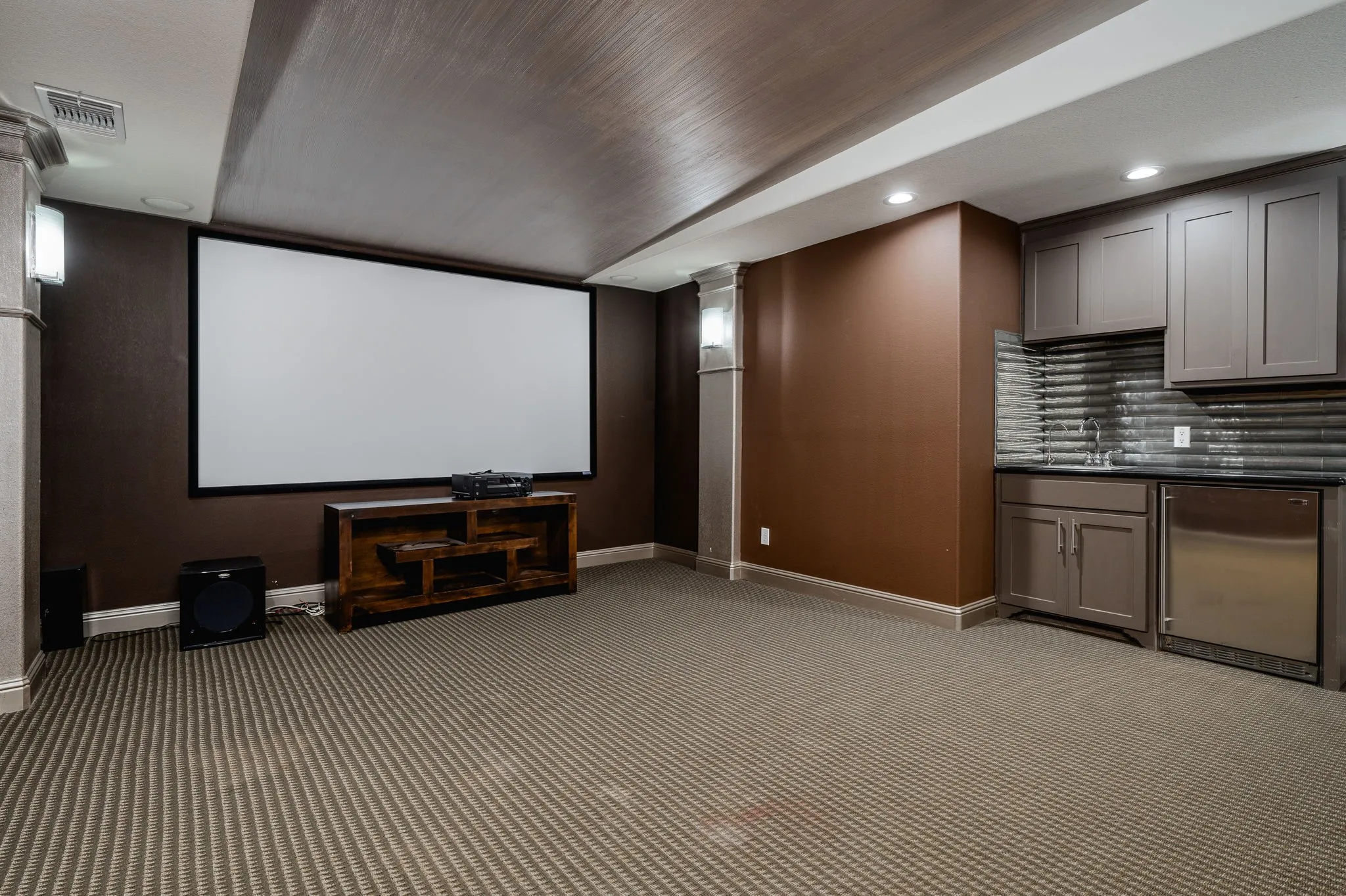 Integrated projection screen, built-in wet bar with cabinetry, stainless steel under-counter refrigerator, textured tile backsplash, and recessed lighting