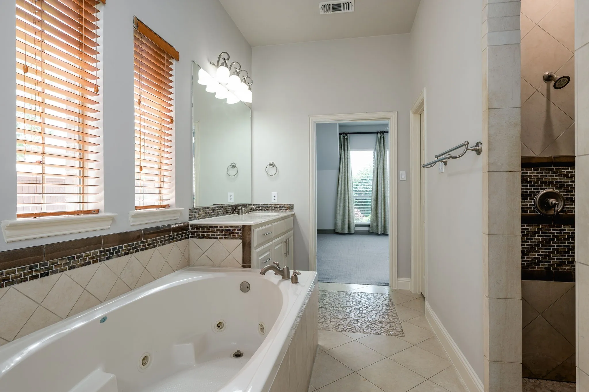 Jetted soaking tub with tiled surround, double vanity with integrated sinks, framed mirror, and a tiled shower with a mosaic accent