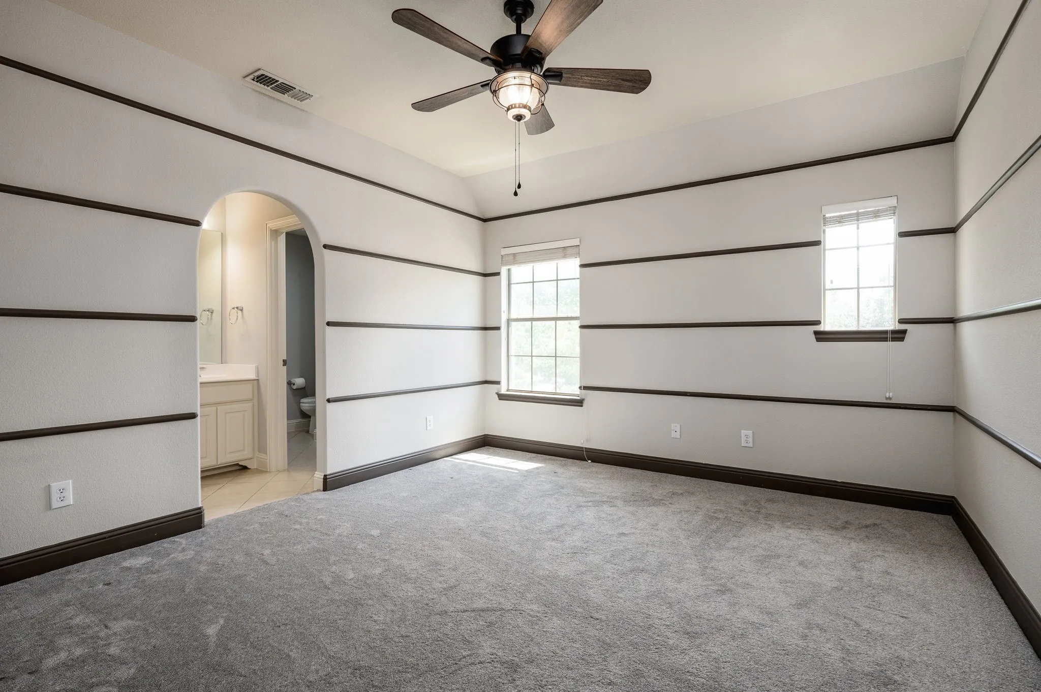 Carpeted room featuring a ceiling fan with light, two windows, and an arched doorway leading to a bathroom with a single vanity