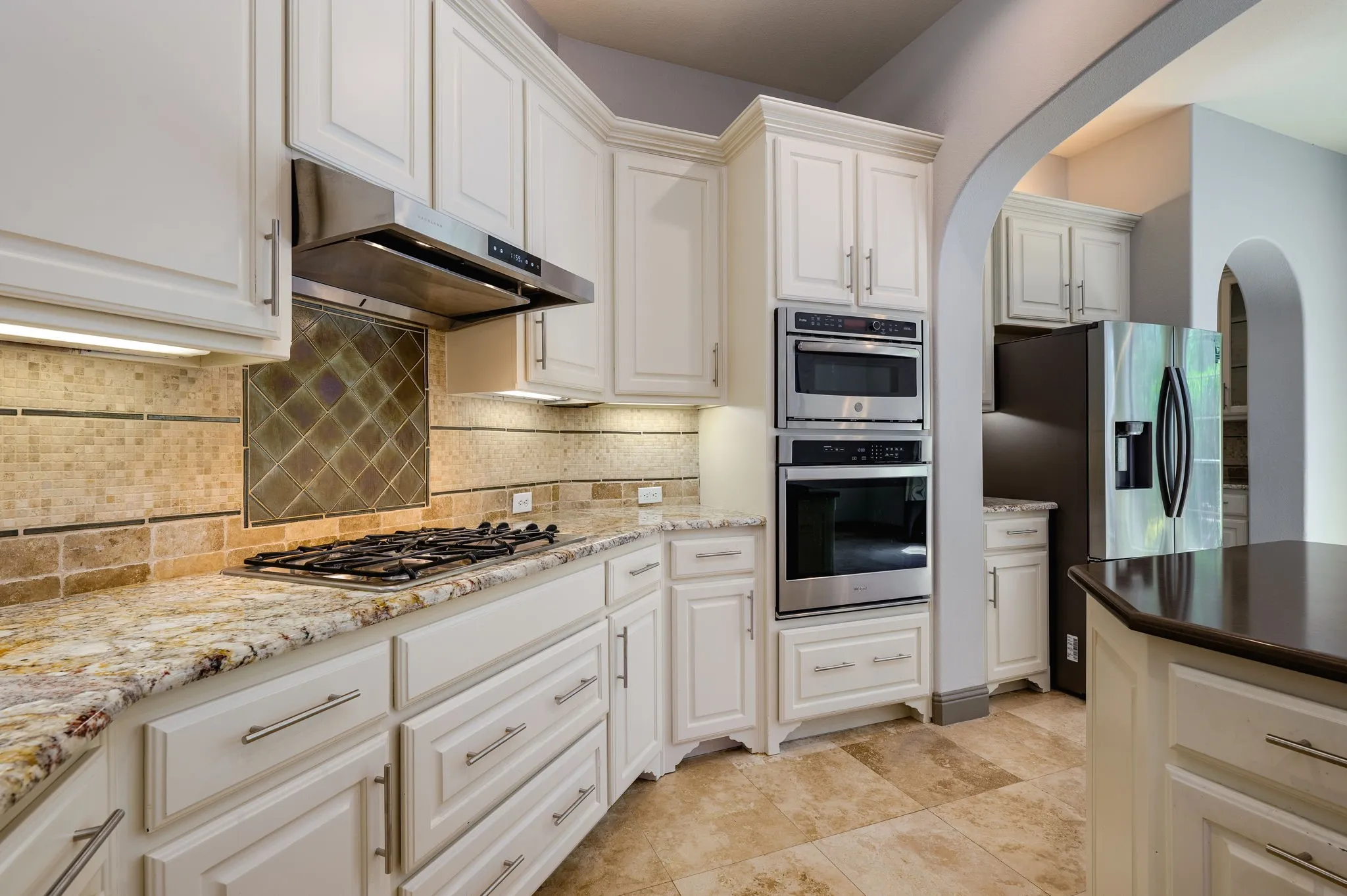 Chef's kitchen featuring cream-raised panel cabinetry, granite countertops, stainless steel dual wall ovens, gas cooktop with decorative tile backsplash, stainless steel range hood, and slate-finish flooring throughout
