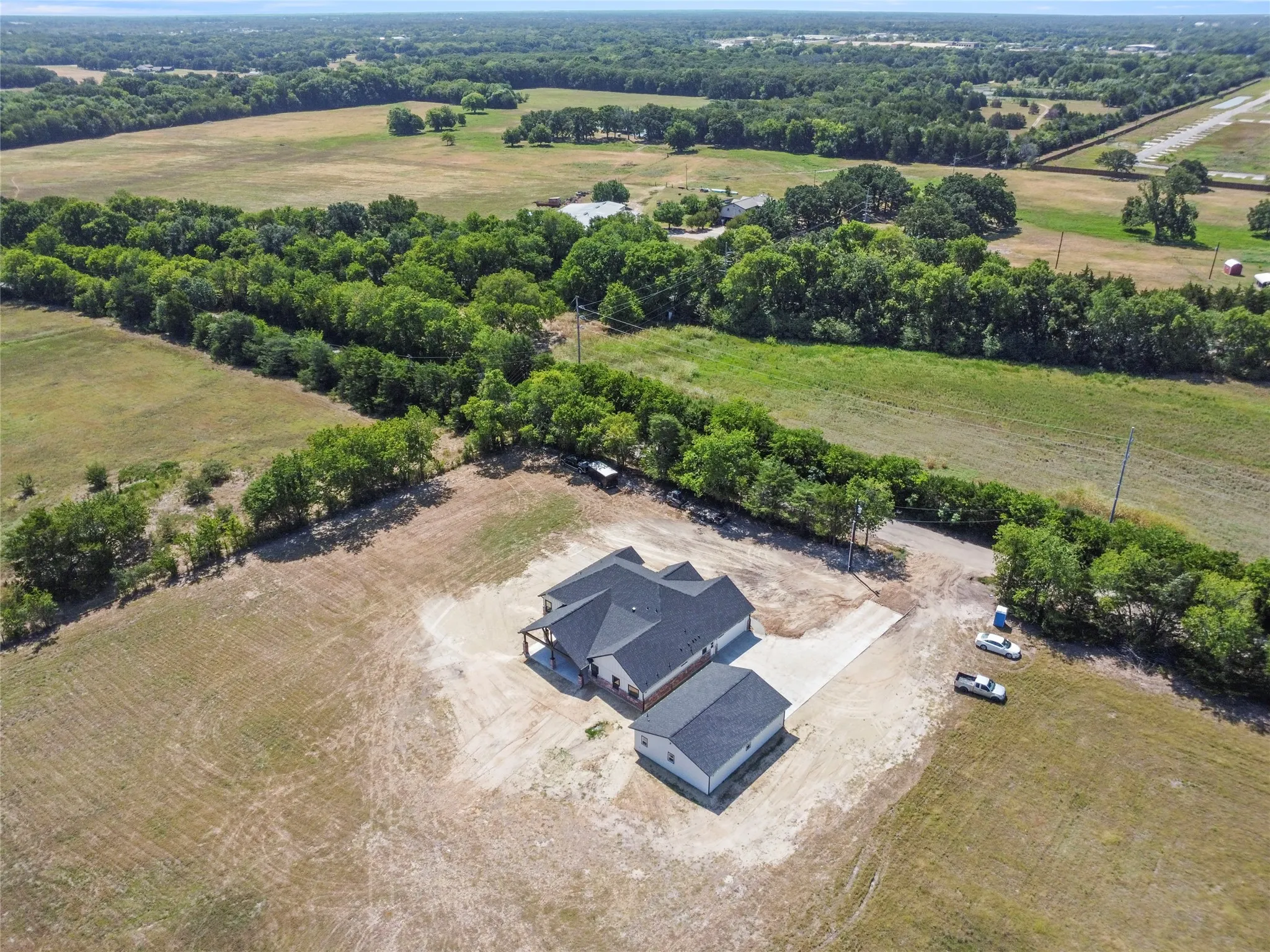 Expansive property featuring a newly constructed residence with an attached garage and a detached accessory structure, surrounded by open fields and mature tree lines