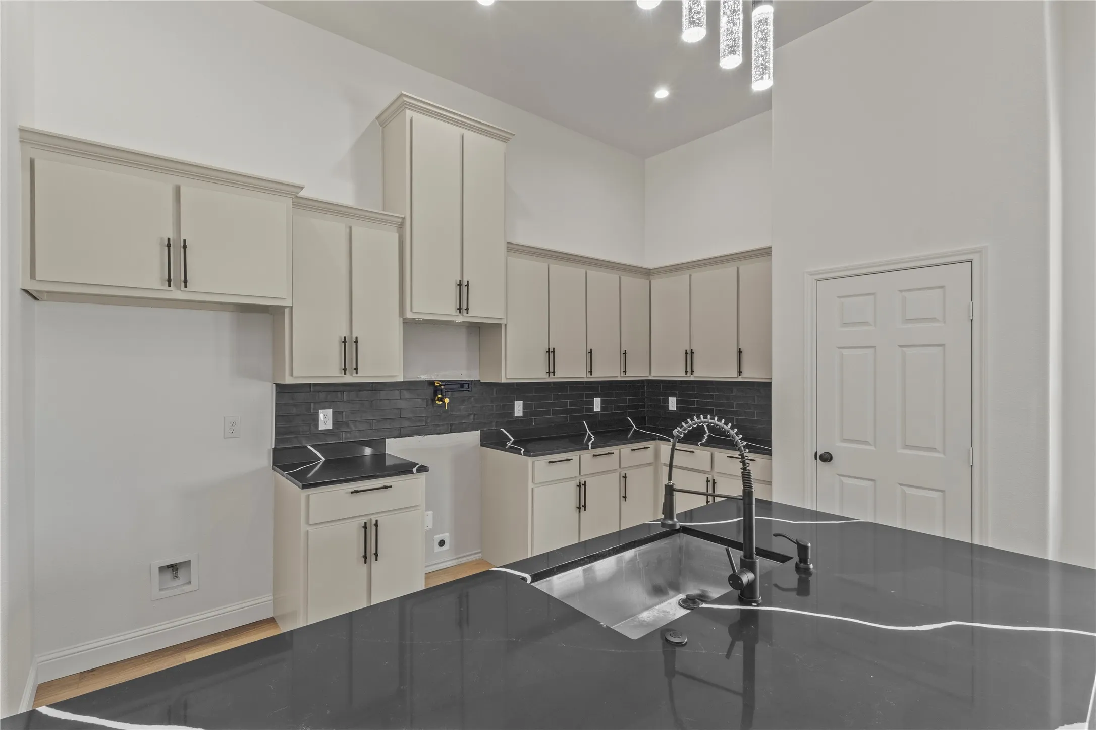 Kitchen featuring light-toned cabinetry, dark subway tile backsplash, dark countertops with light veining, wood-finish flooring, and an island with an integrated sink and faucet