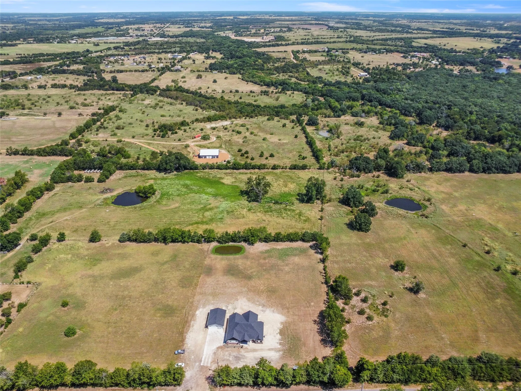 Expansive acreage featuring multiple ponds, mature trees, a modern residence with a dark roof, and a separate outbuilding