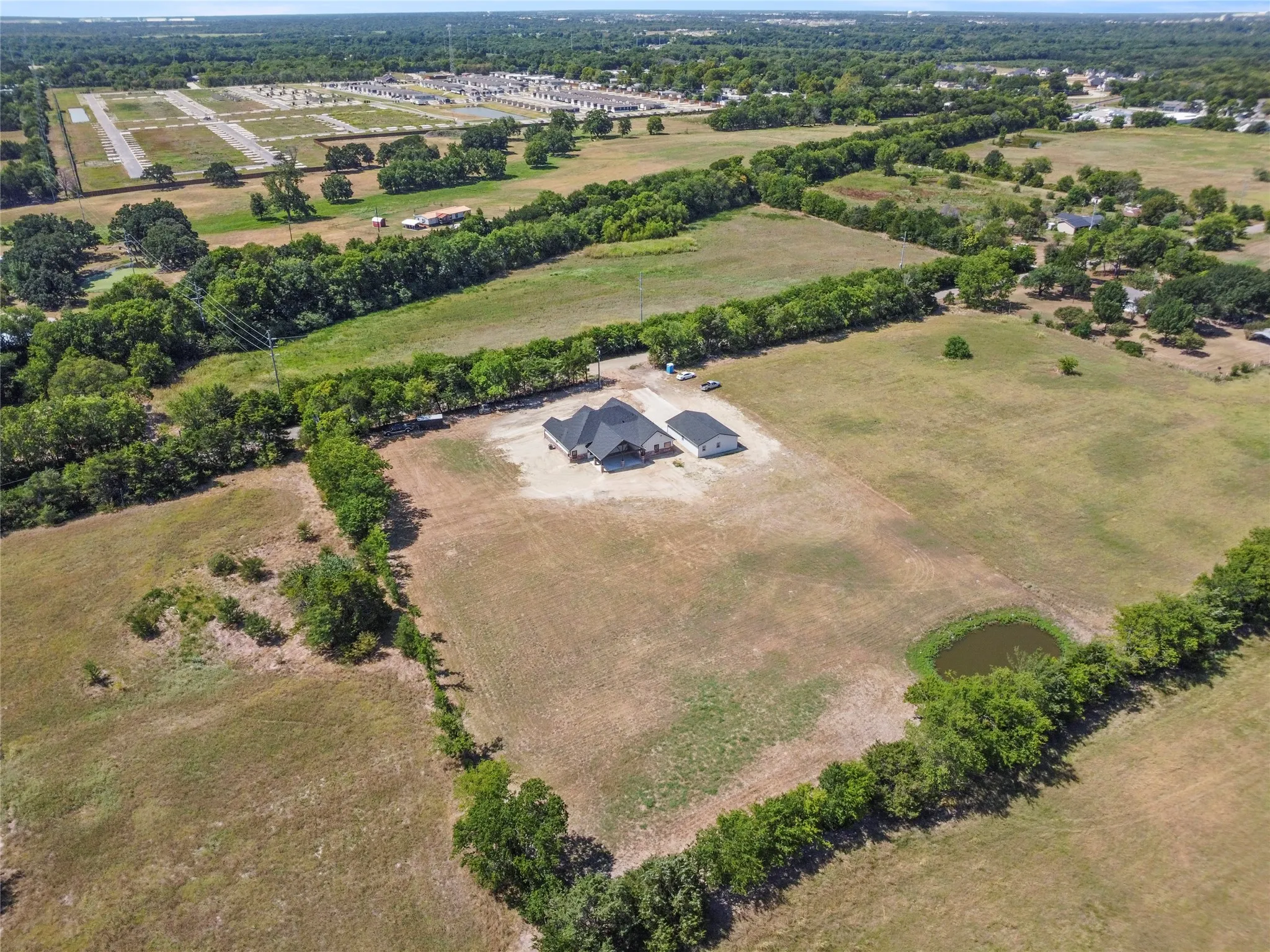 Expansive property featuring a single-story residence with a dark roof, an accessory building, surrounding fields, and a small pond