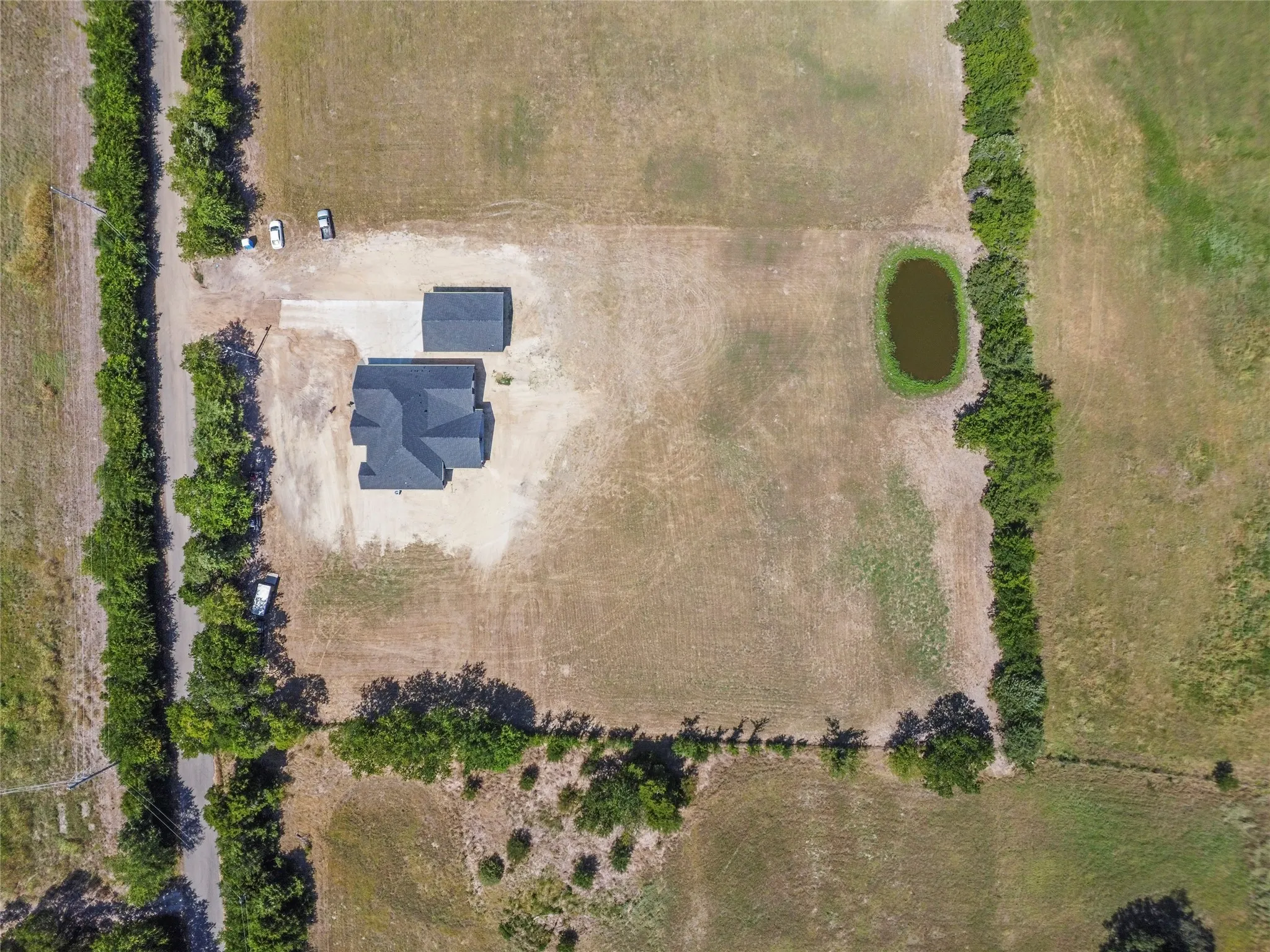 Expansive property featuring a residence with a dark roof, a detached structure with a matching roof, a gravel driveway, and a natural pond