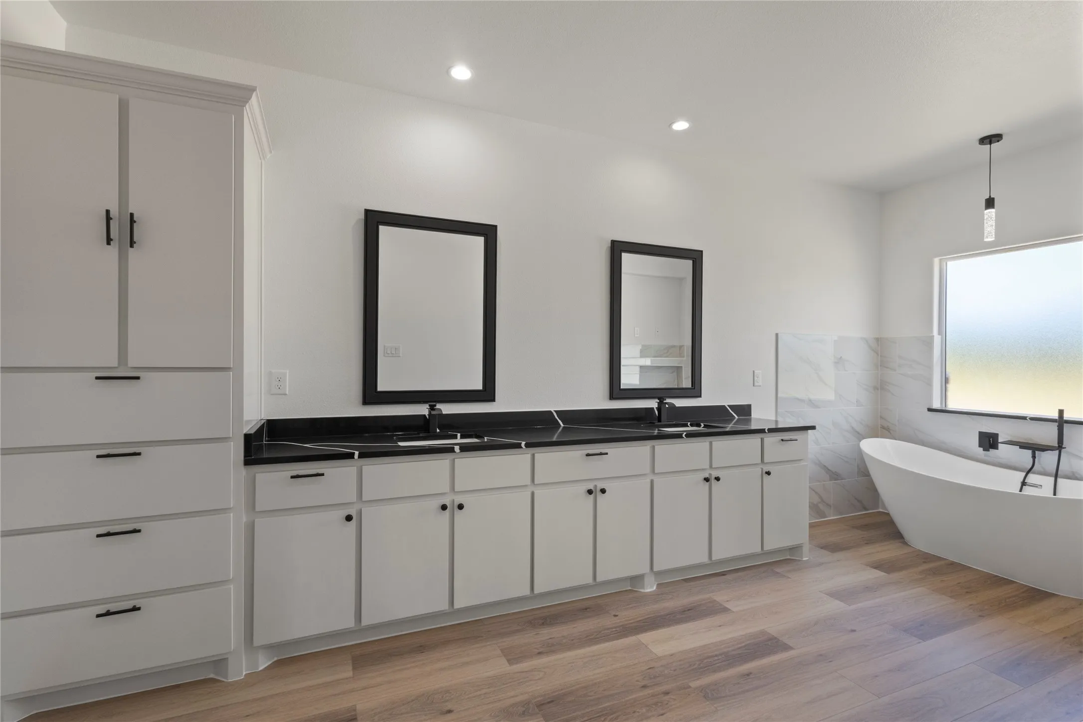 Spa-inspired primary bathroom featuring a freestanding soaking tub, dual vanity with black countertops and integrated sink basins, wood-finish flooring, recessed ceiling lighting, and large framed mirrors with black trim