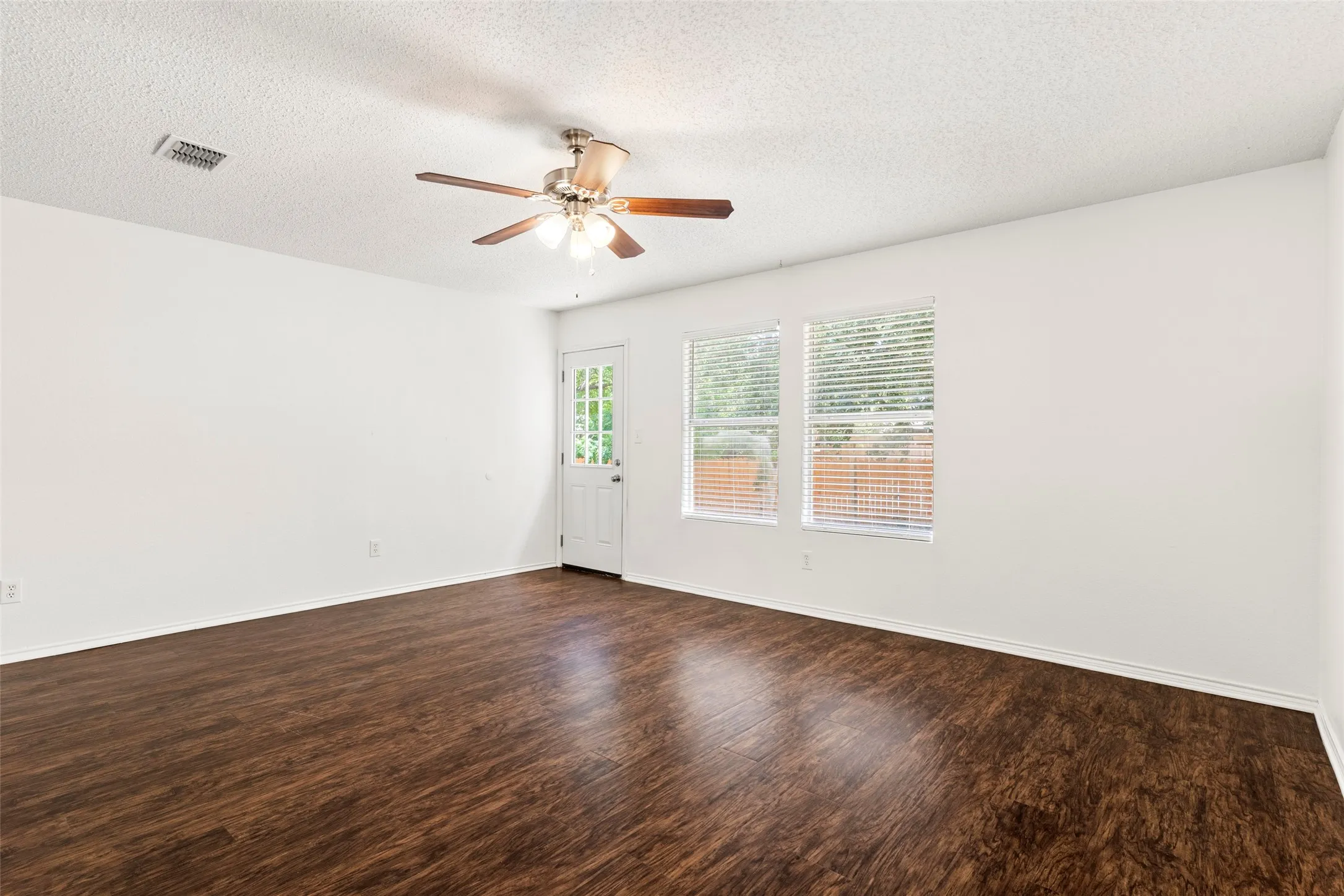 Wood-finish flooring, ceiling fan with light fixture, white interior walls, two windows with blinds, and an exterior door with glass panels