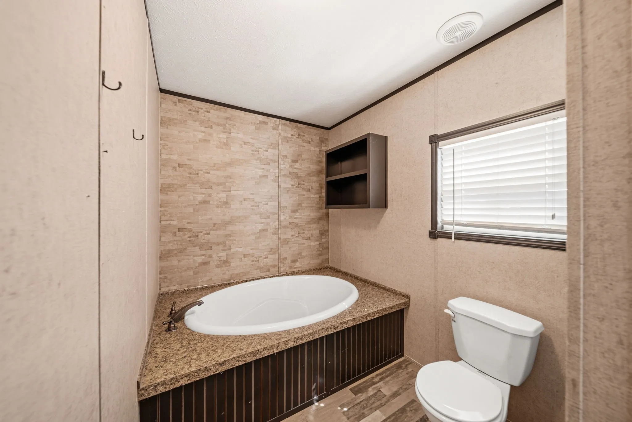 Corner soaking tub with stone tile surround, granite-look countertop, and built-in shelving