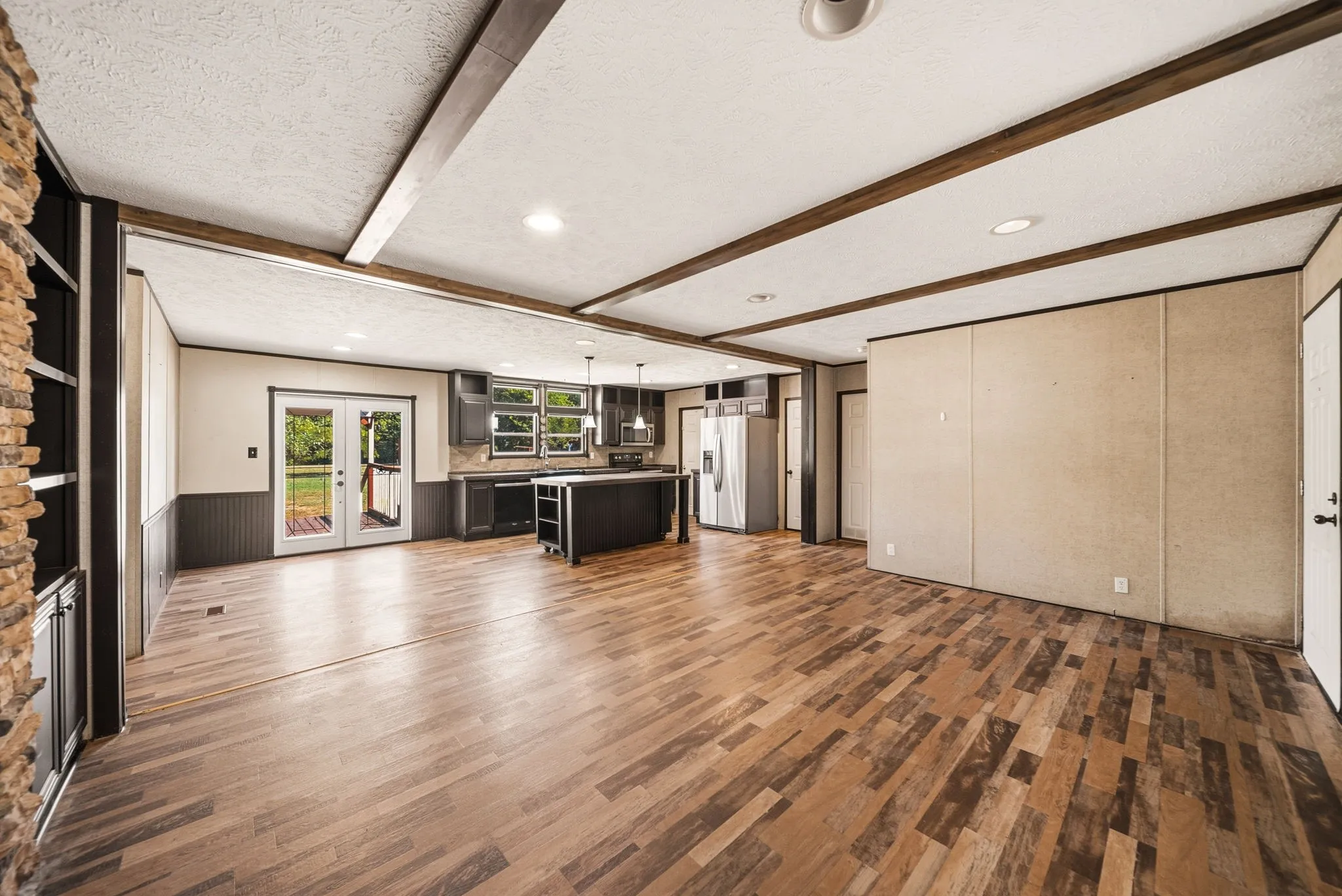 Open concept interior featuring wood-finish flooring, exposed ceiling beams, a stone-faced built-in shelving unit, and a kitchen with dark cabinetry and an island