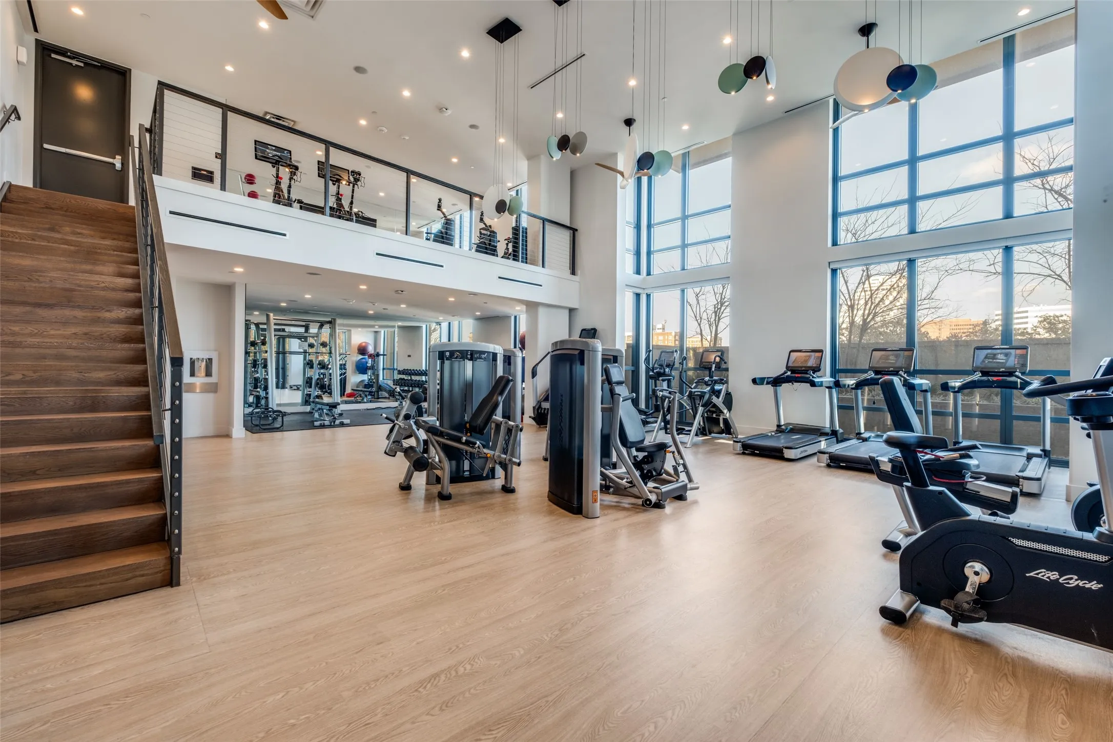Two-story fitness center featuring wood-finish flooring, a wood staircase with metal railings, and extensive natural light from large windows