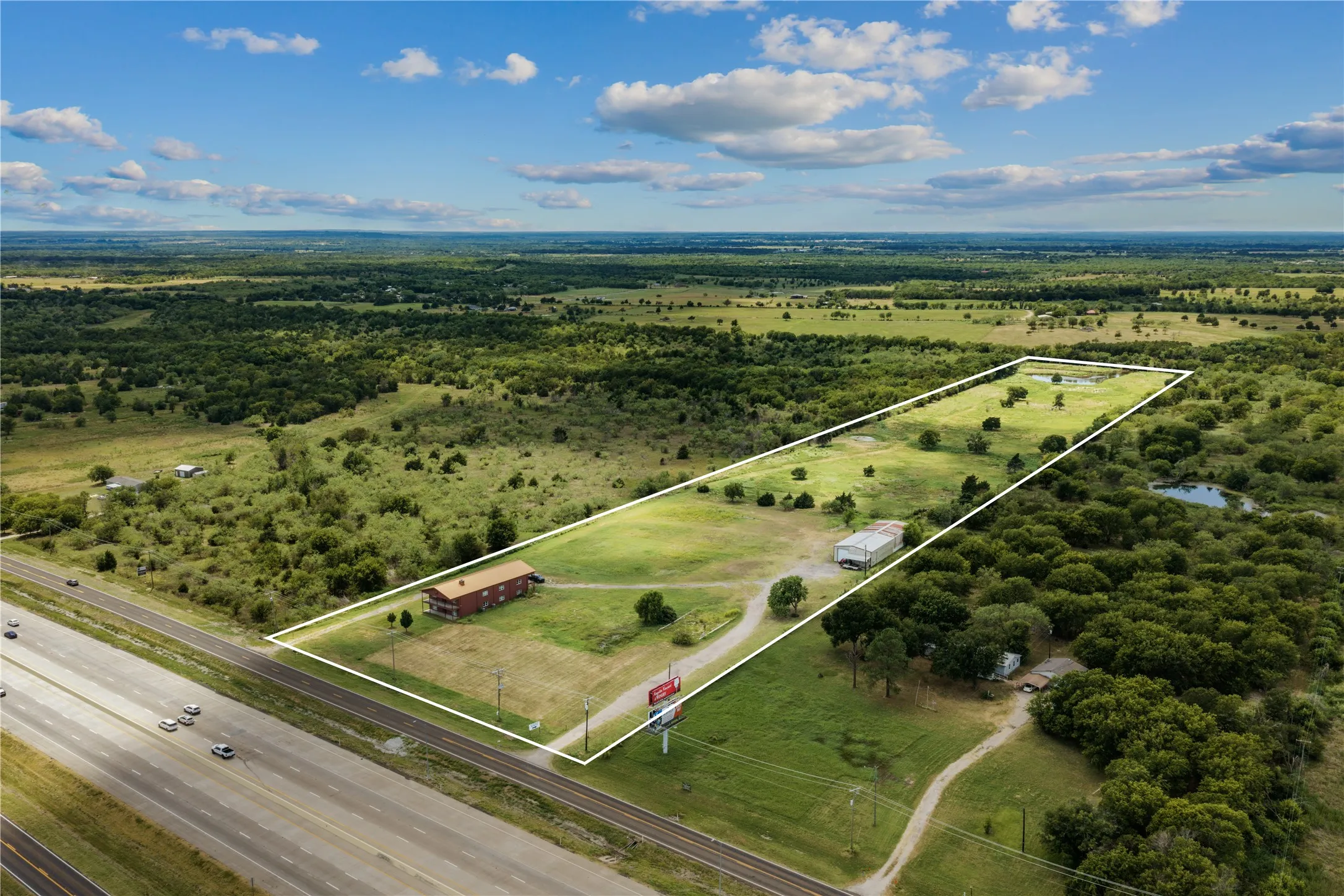 Expansive acreage featuring a red-roofed building, a large outbuilding, a driveway, and multiple ponds, bordered by a highway and natural landscape