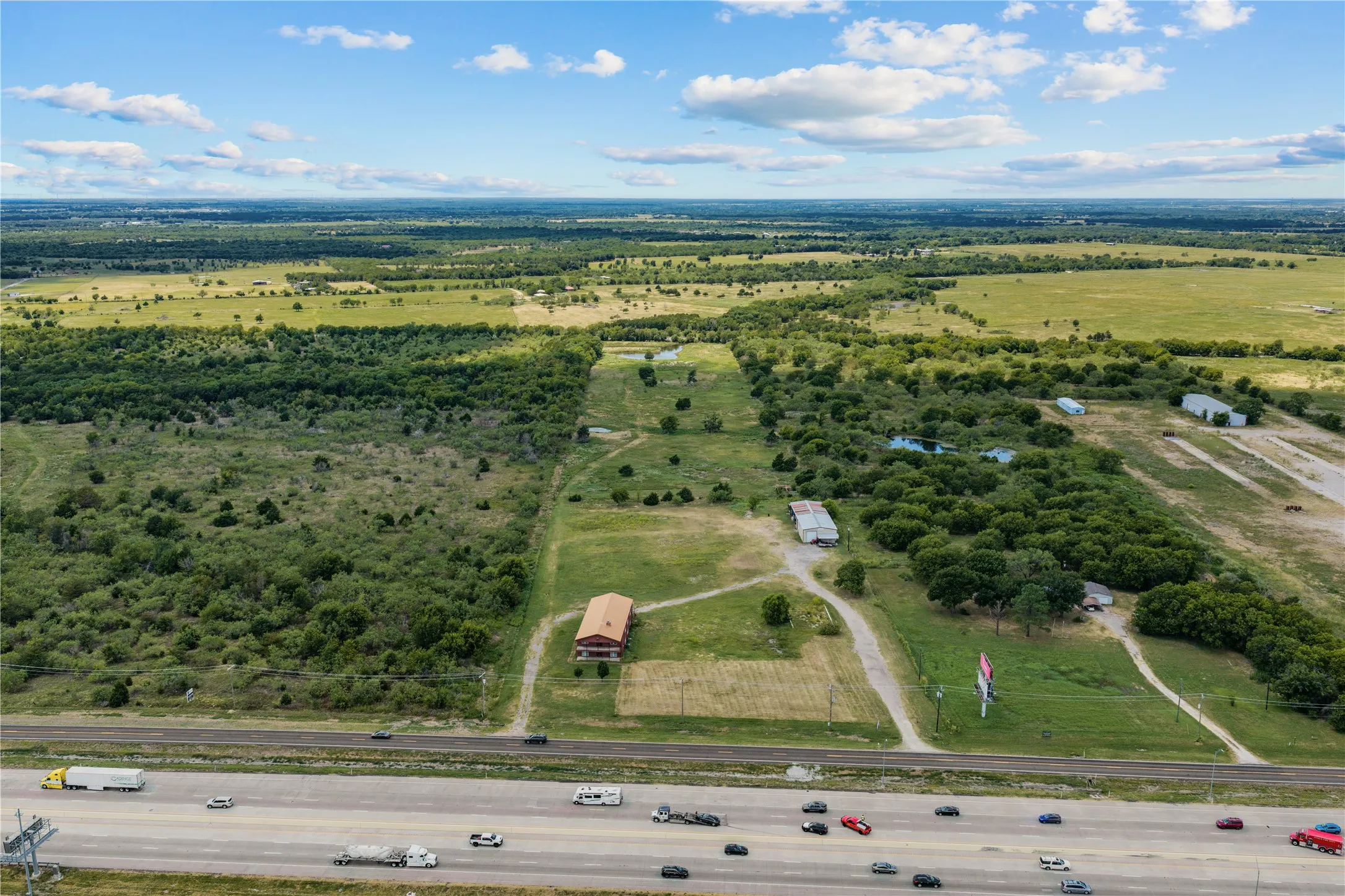 Expansive undeveloped land with a significant tree line, multiple outbuildings, and a large red-roofed structure