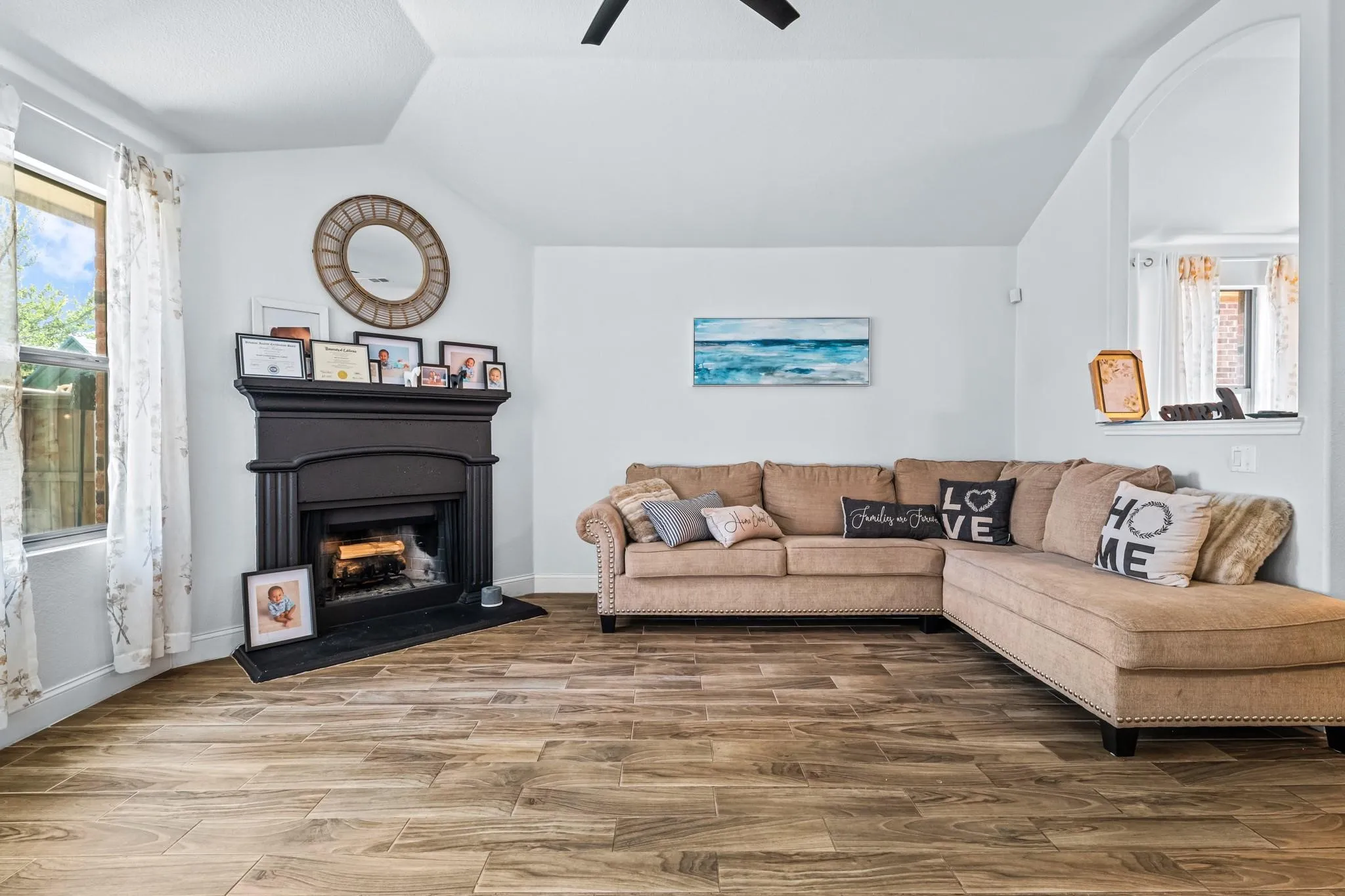 Fireplace with dark mantel, wood-finish flooring, window with natural light, and a ceiling fan