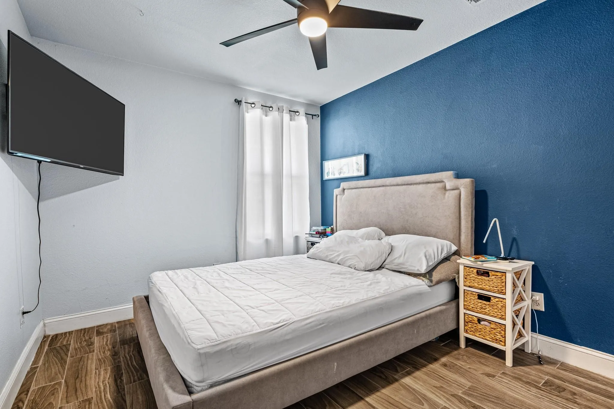 Wood-finish flooring, accent wall, ceiling fan, and window with privacy curtains