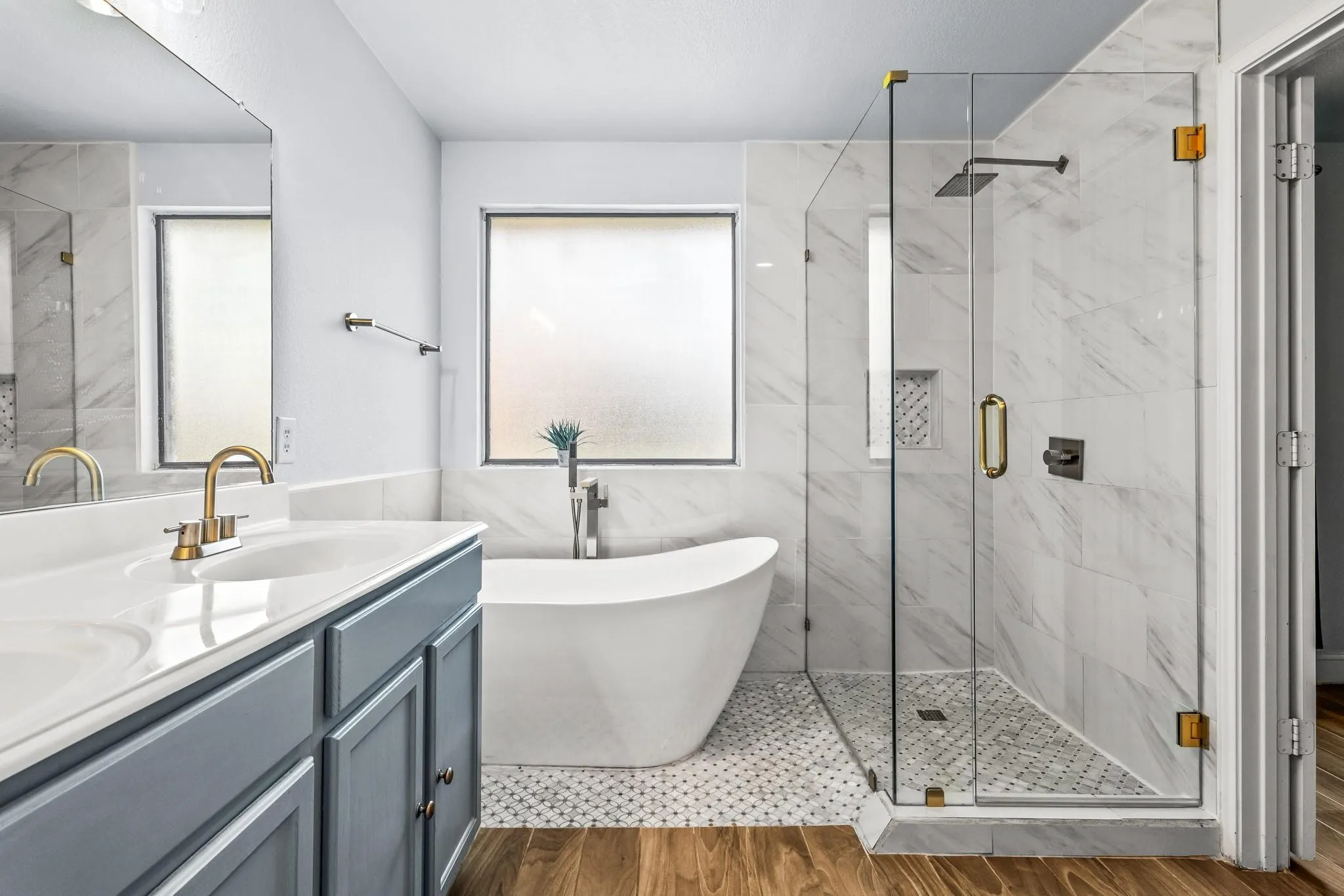 Freestanding soaking tub, glass-enclosed shower with rainhead, double vanity with gray cabinetry, and wood-finish flooring