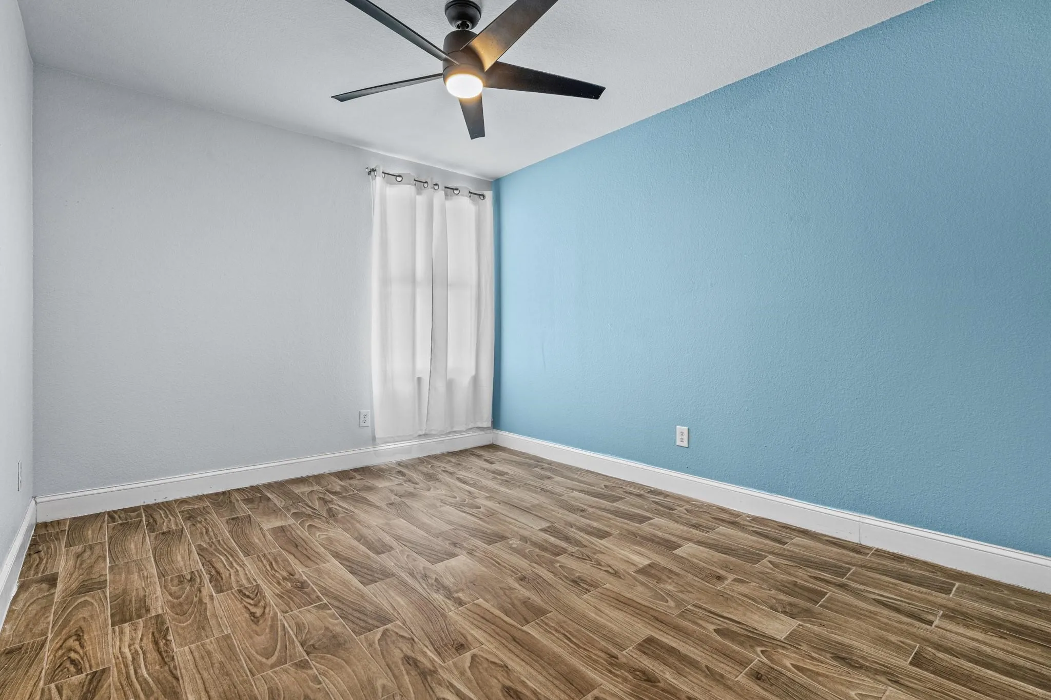 Wood-finish flooring, ceiling fan with integrated lighting, window with white curtains, and a two-tone wall finish